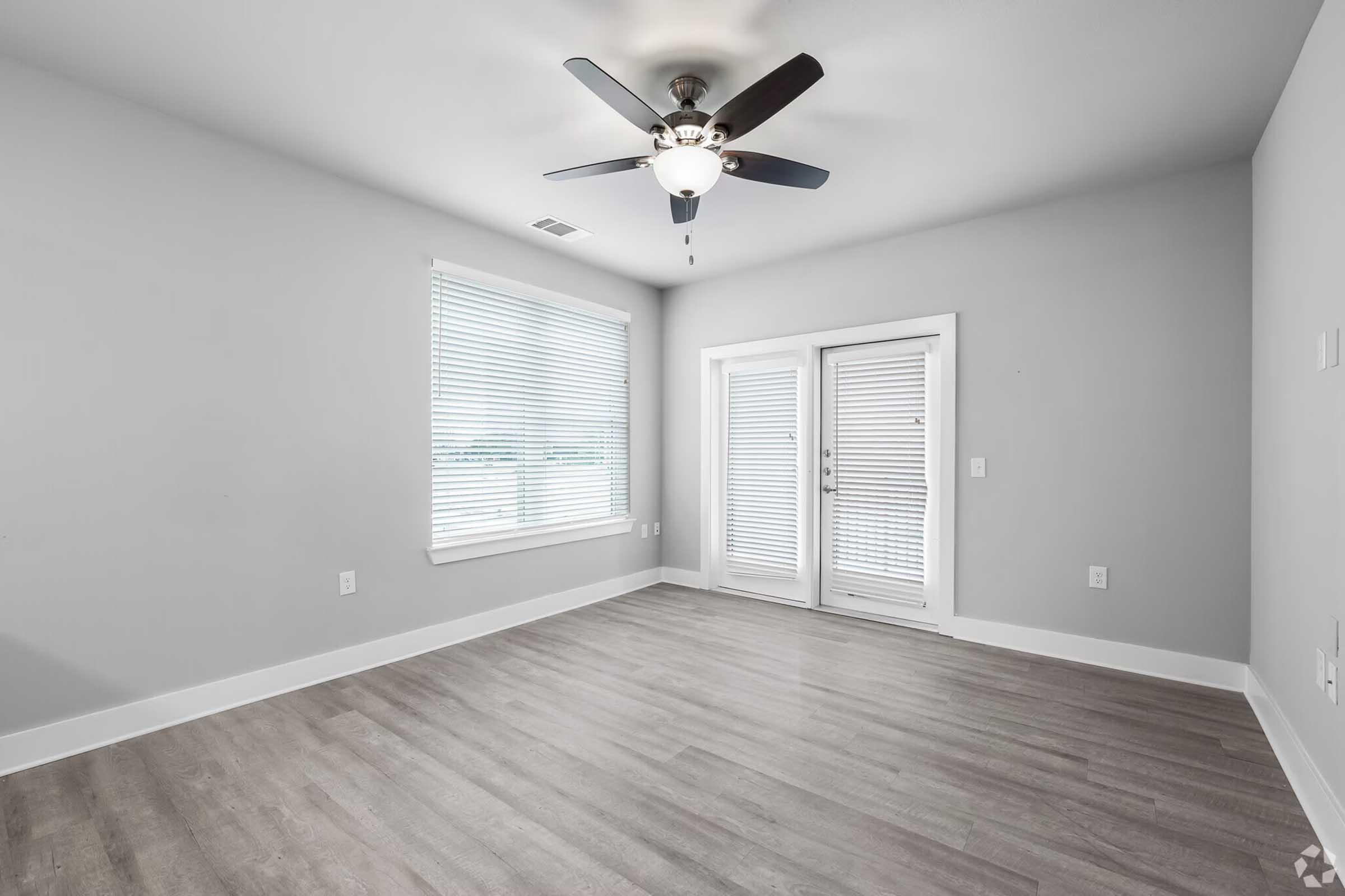 Empty room with gray walls, a ceiling fan, and light-colored wooden flooring. Features large windows with blinds and a set of double doors leading outside. The space is uncluttered and well-lit, creating a modern and airy atmosphere.