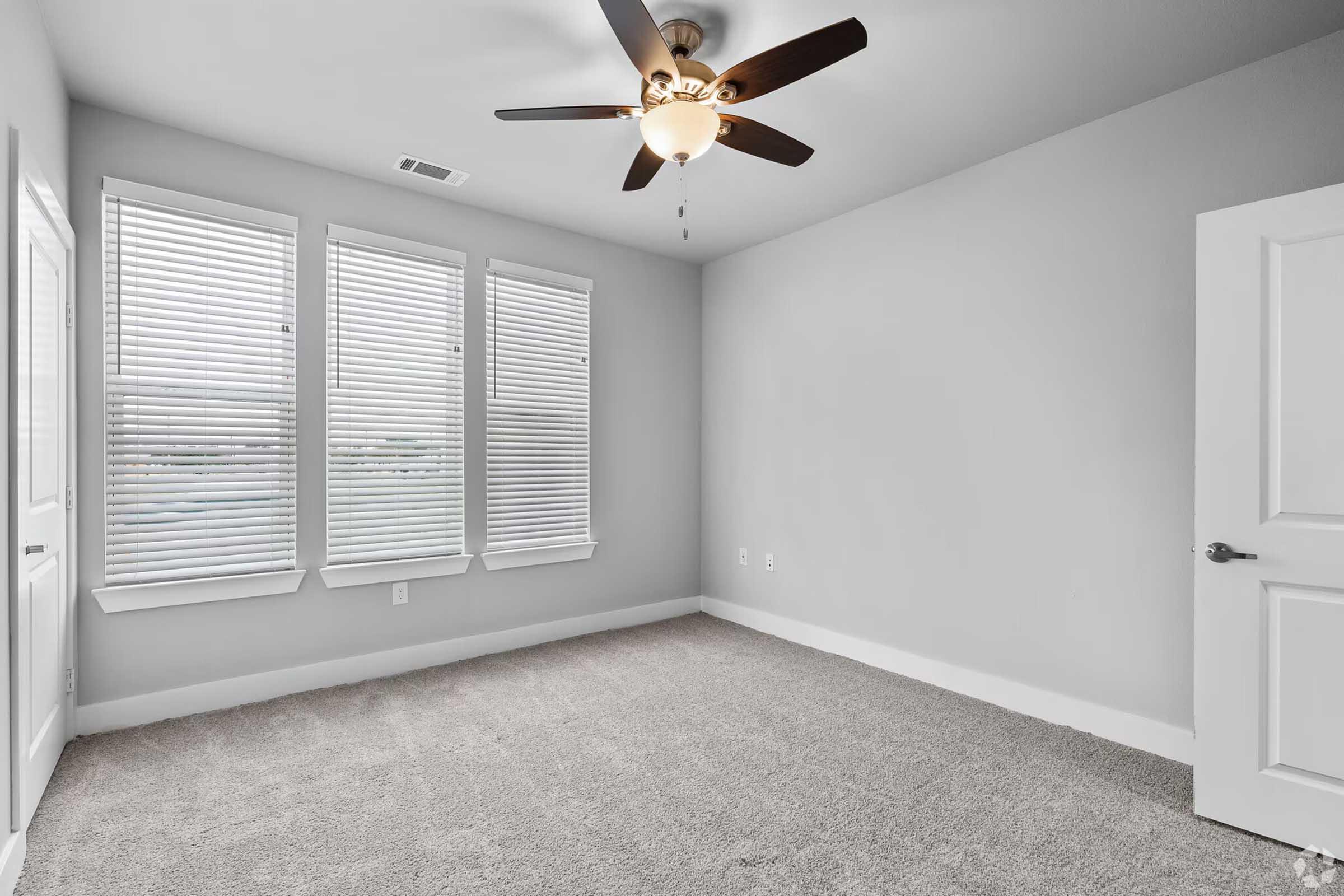 A spacious, empty room with light gray walls and carpeted flooring. It features three large windows with white blinds allowing natural light, and a ceiling fan with a light fixture in the center. The door is closed, and there are no furnishings present, creating a minimalist atmosphere.