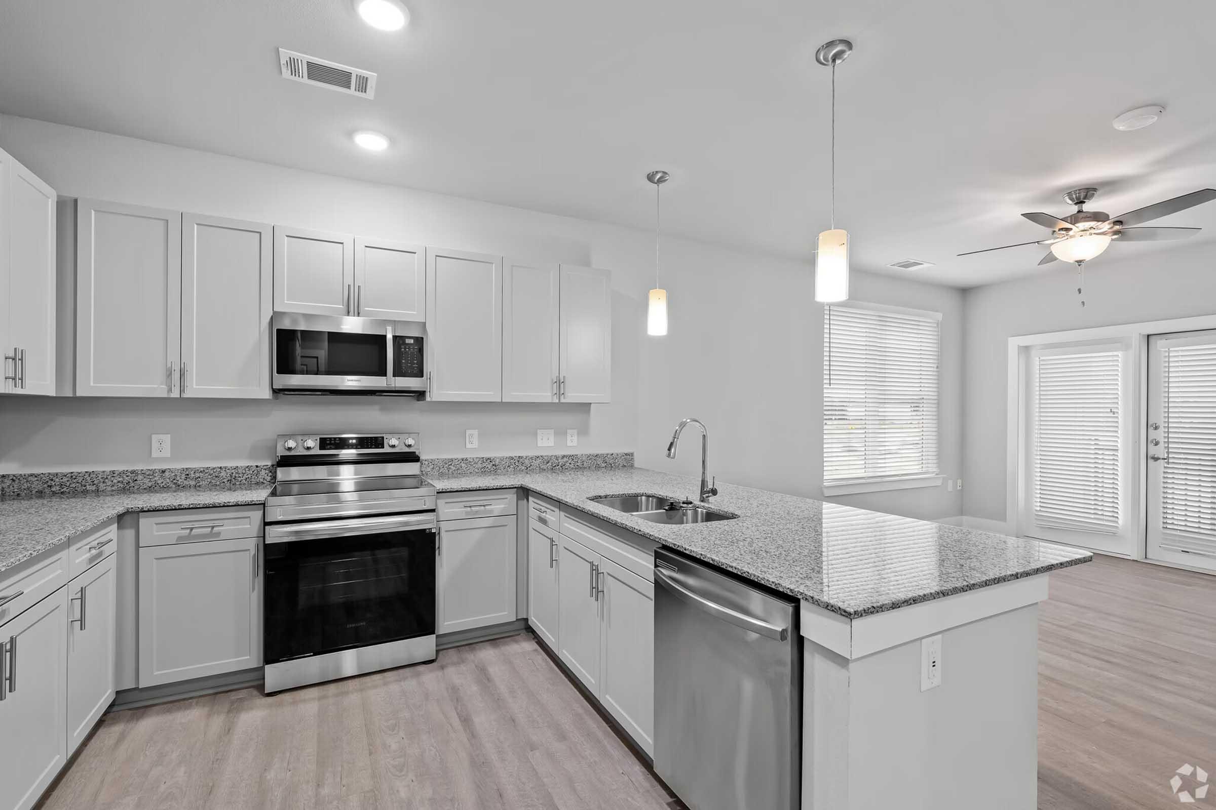 Modern kitchen featuring stainless steel appliances, including a microwave and oven. The island includes a sink, and there are light gray cabinets with granite countertops. Bright, well-lit space with large windows, a ceiling fan, and nearby sliding glass doors leading to an outdoor area.