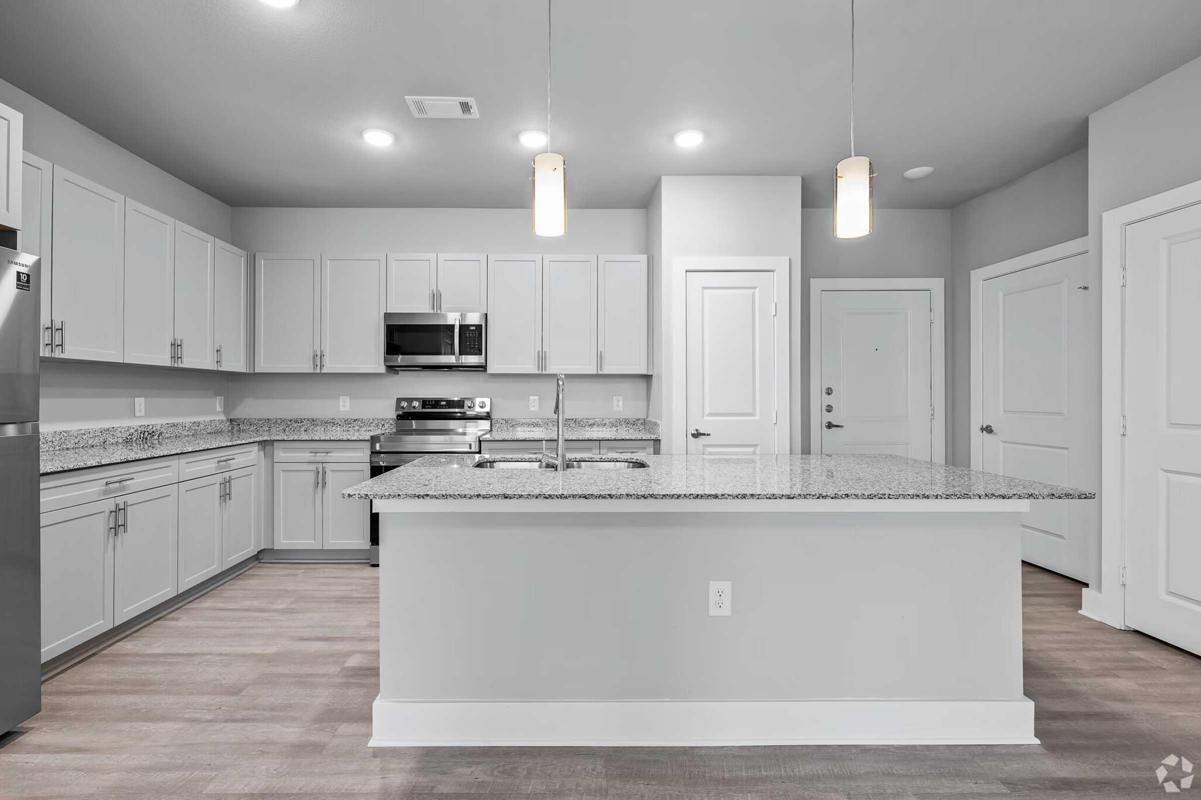 Modern kitchen with gray cabinets, stainless steel appliances, and granite countertops. There's an island with seating space and pendant lighting above. The walls are light gray, and the flooring is a wood-like laminate. Two doors lead to a hallway and the entrance.