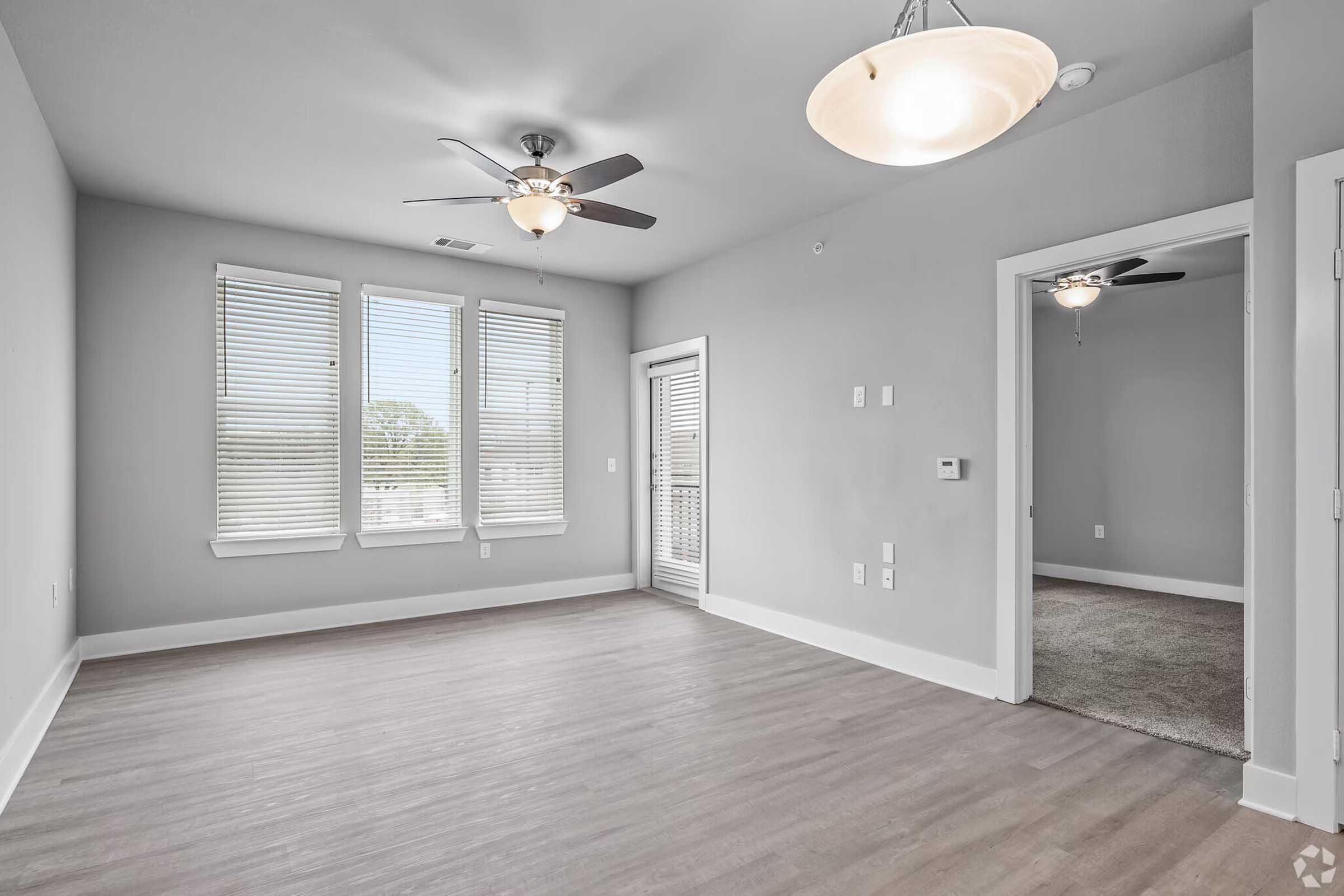 A spacious room with light gray walls, large windows with white blinds, and a ceiling fan. There is a doorway leading to another room and a sliding glass door that opens to a balcony. The flooring is light-colored wood, and the room is well-lit with natural light.