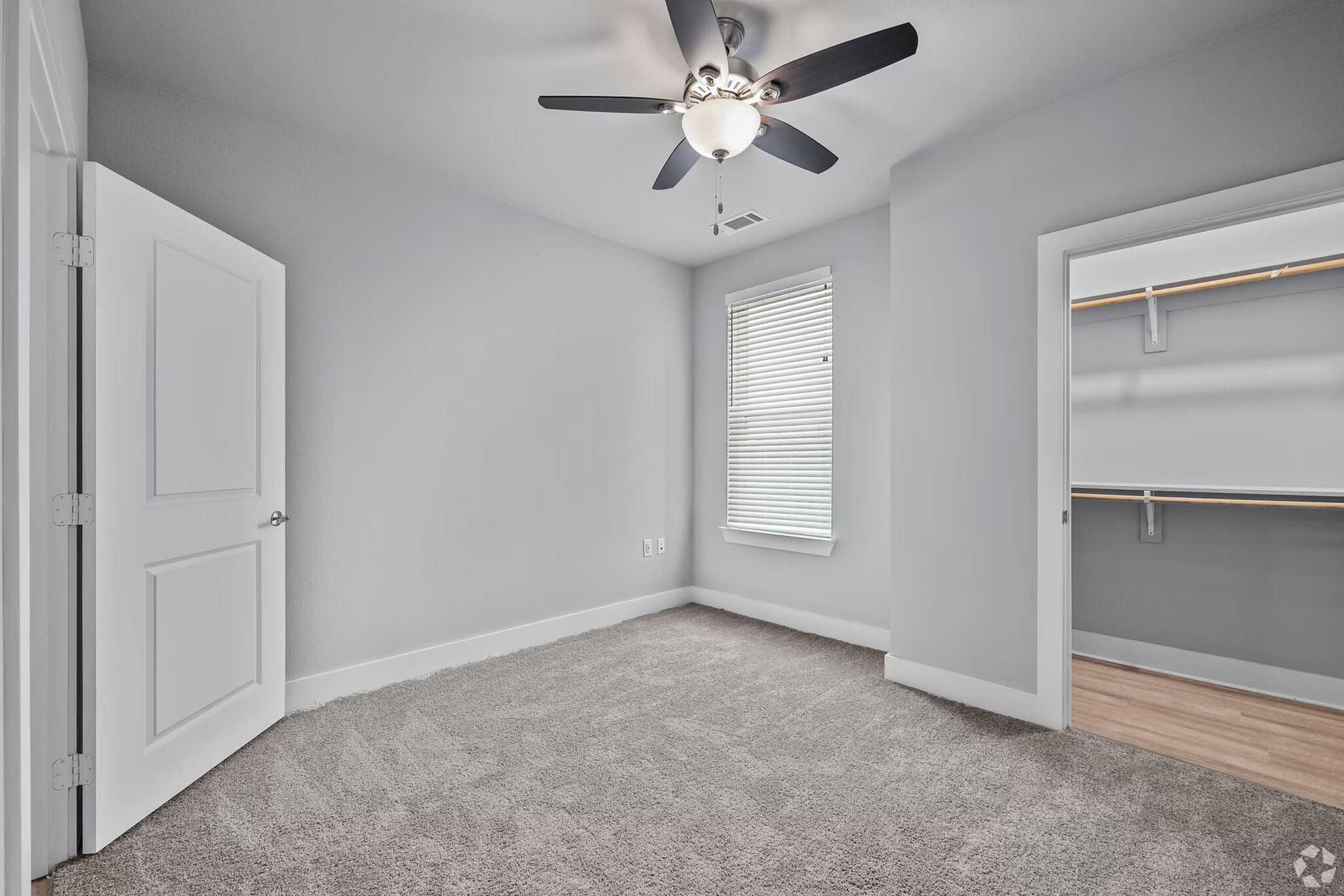 A spacious, empty bedroom featuring light gray walls and plush carpet. The room includes a ceiling fan, a window with blinds allowing natural light, and an open closet with shelving. A door on the left leads to another area, enhancing the room's accessibility.