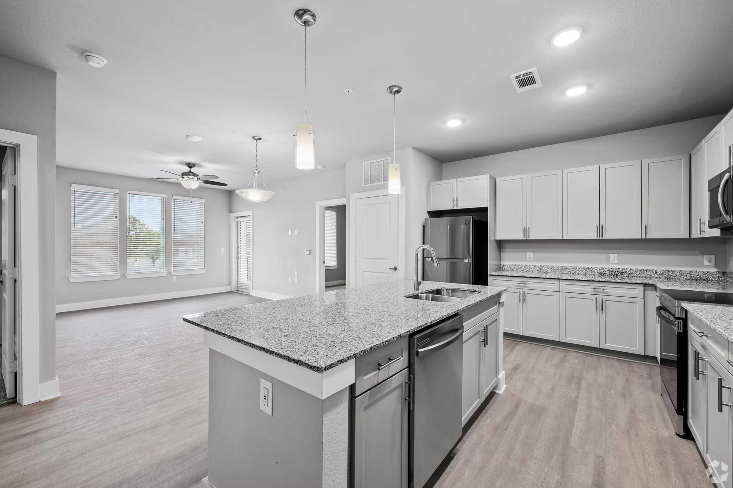 Modern kitchen with granite countertops, stainless steel appliances, and a spacious island. The open layout connects to a bright living area with large windows and light-colored walls. Features include pendant lighting and wood-style flooring.