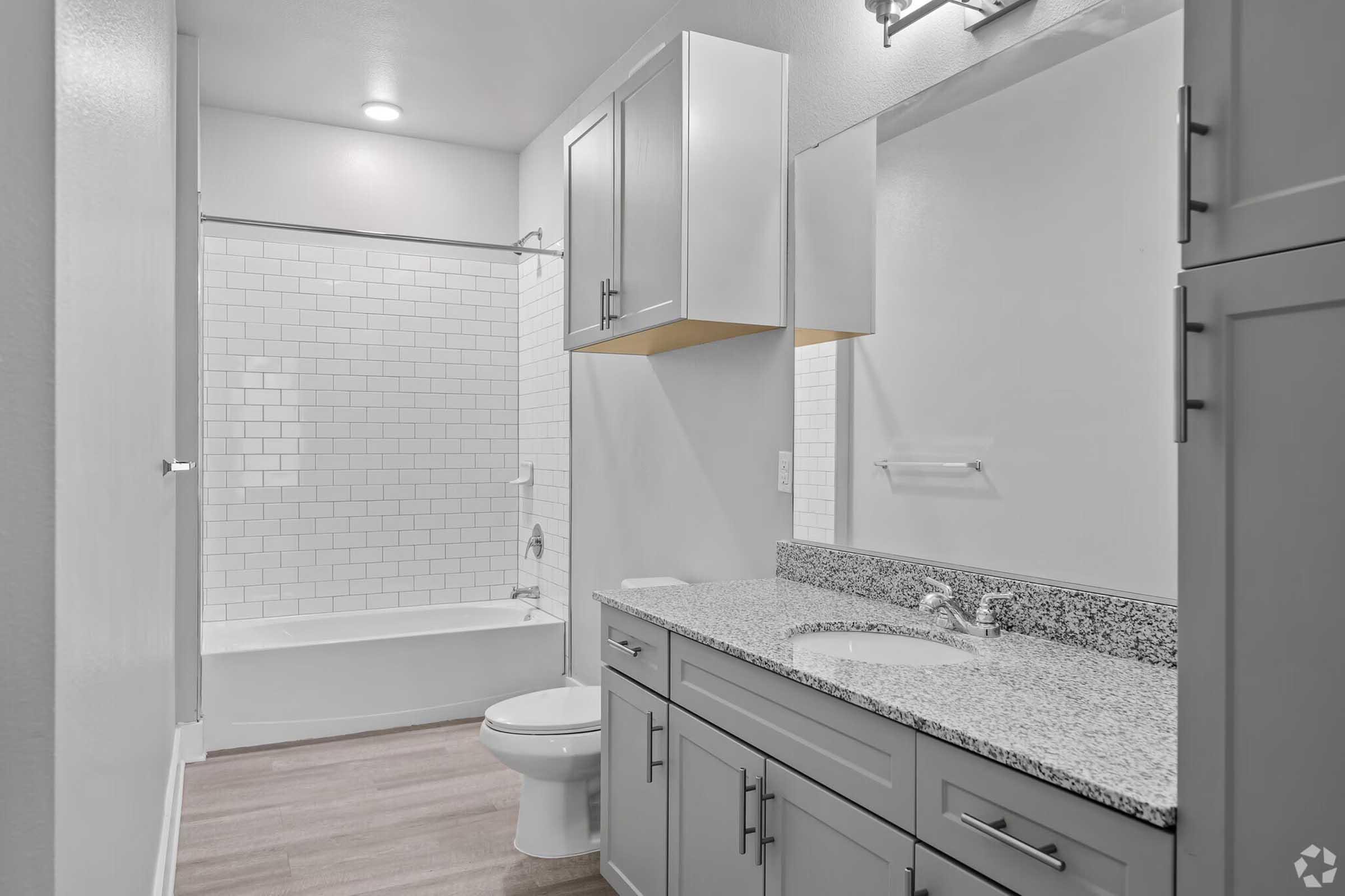 Modern bathroom featuring a white tiled shower with a bathtub, a double vanity with a speckled countertop, and light gray cabinets. The walls are painted in a neutral tone, and there is overhead lighting above the mirror. The flooring is light wood, enhancing the contemporary look of the space.