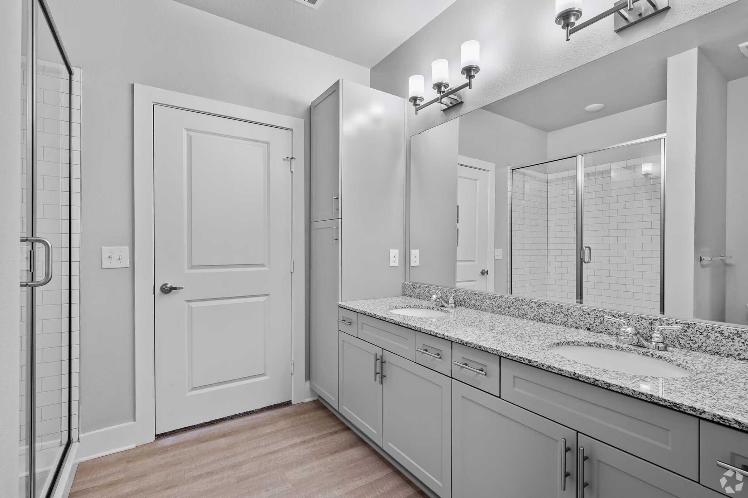 Modern bathroom featuring a double vanity with two sinks, granite countertop, and ample storage. A walk-in shower with glass doors is visible, along with a closed door leading to another room. Bright lighting from wall-mounted fixtures illuminates the space, complemented by neutral-colored walls and flooring.