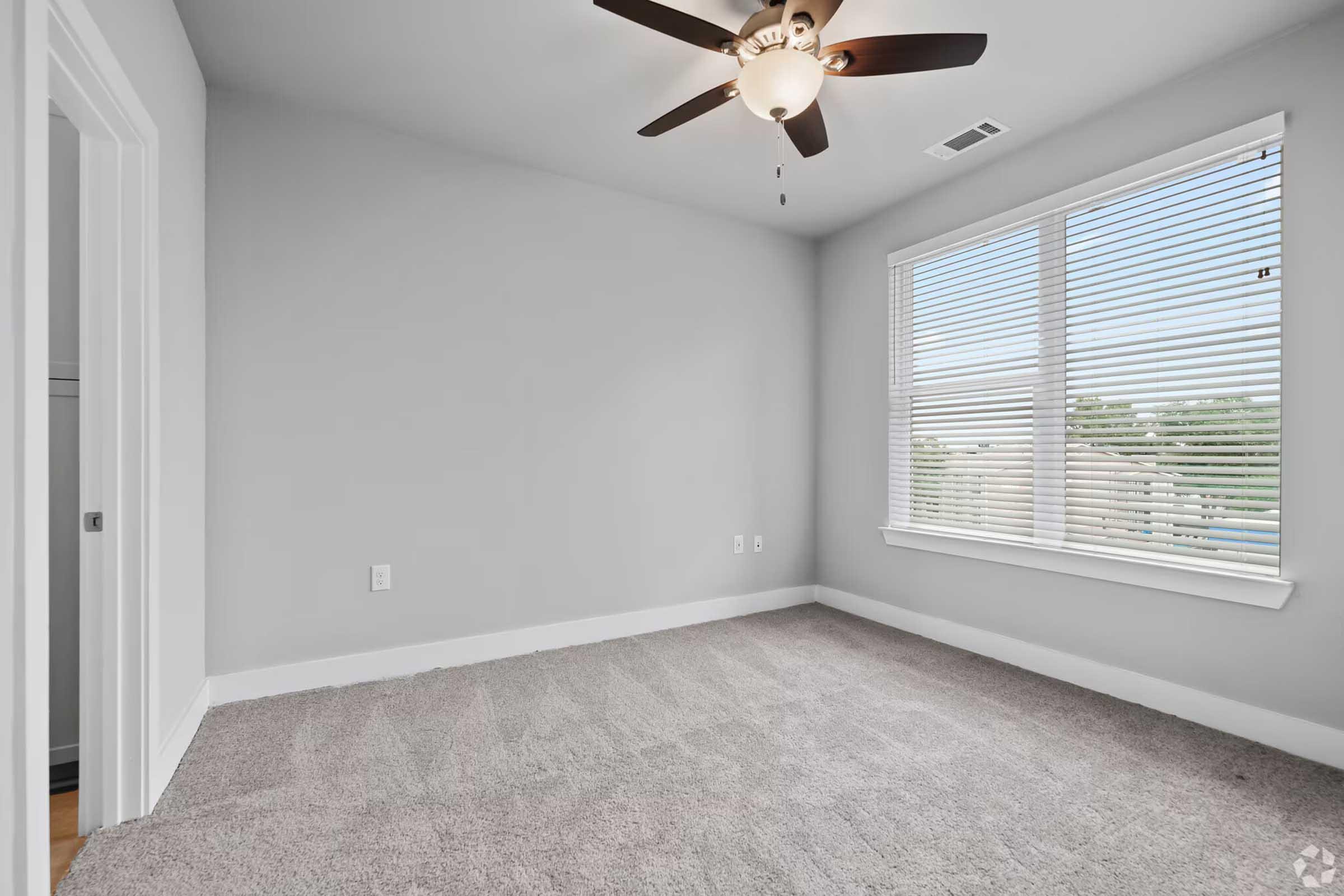A well-lit, empty room featuring light gray walls and a carpeted floor. There is a ceiling fan with wooden blades and a window dressed with blinds, allowing natural light to enter. A doorway on the left leads to another space, while the room has a minimalist and spacious feel.
