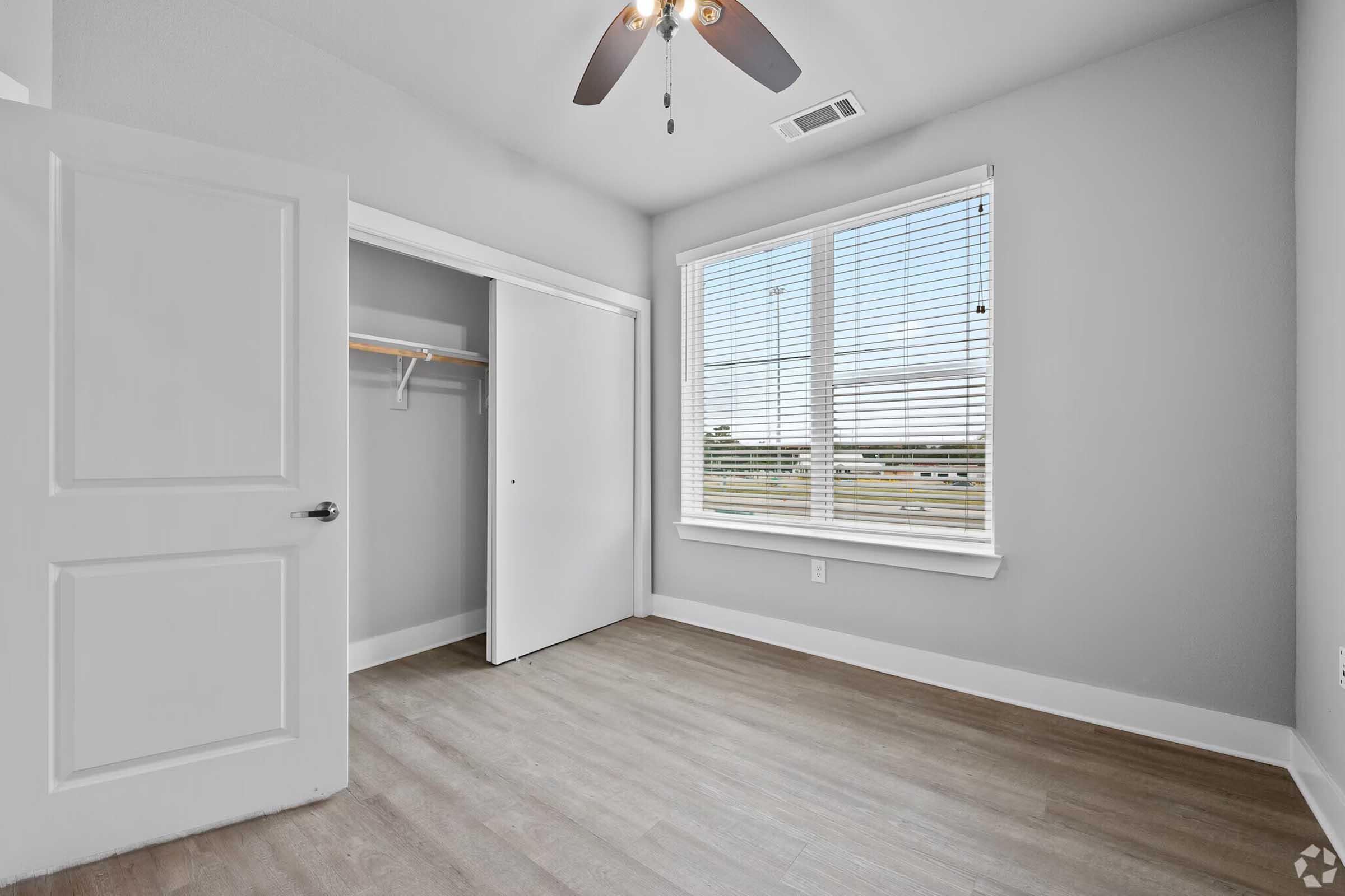 Bright room with light gray walls and a ceiling fan. A window with blinds provides natural light, and there's a closet with a sliding door and a shelf inside. The flooring is light-colored wood laminate, creating a clean and modern atmosphere.