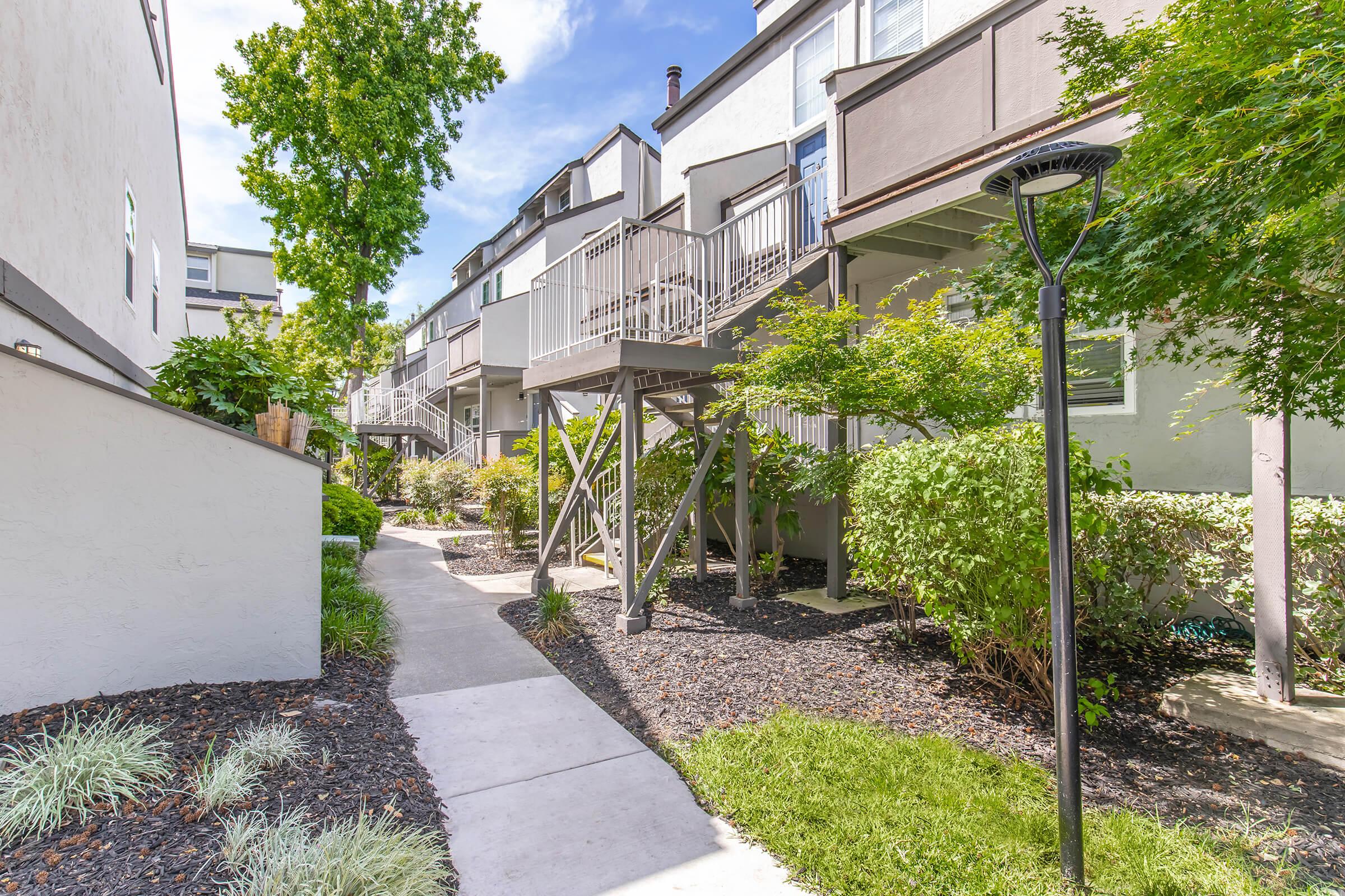 A well-maintained pathway surrounded by green shrubs and small trees, leading through a modern residential area. The buildings have multiple levels with balconies, and a lamp post stands alongside the path, creating a pleasant, inviting atmosphere. Sunlight filters through the trees on a clear day.
