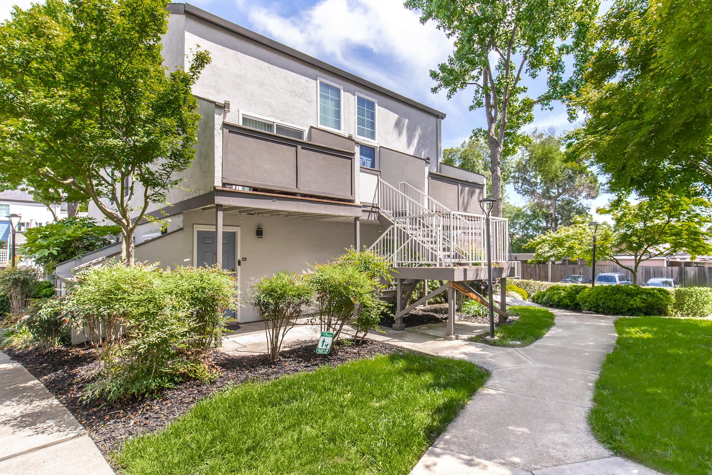A modern two-story building with a light gray exterior, featuring a staircase leading to a second-floor entrance. Surrounding the structure are lush green trees and shrubs, with a walkway and manicured lawn. Soft sunlight illuminates the scene, creating a welcoming atmosphere.