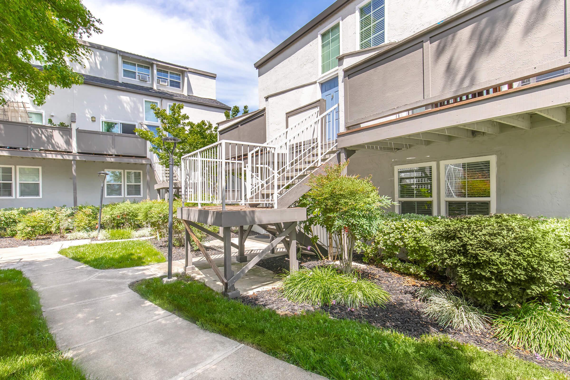 A residential building with multiple stories, featuring a staircase leading to a second-floor entrance. The area is landscaped with green grass and shrubs, and there are walking paths and outdoor lighting fixtures. The sky is clear with a few clouds, contributing to a bright and inviting atmosphere.