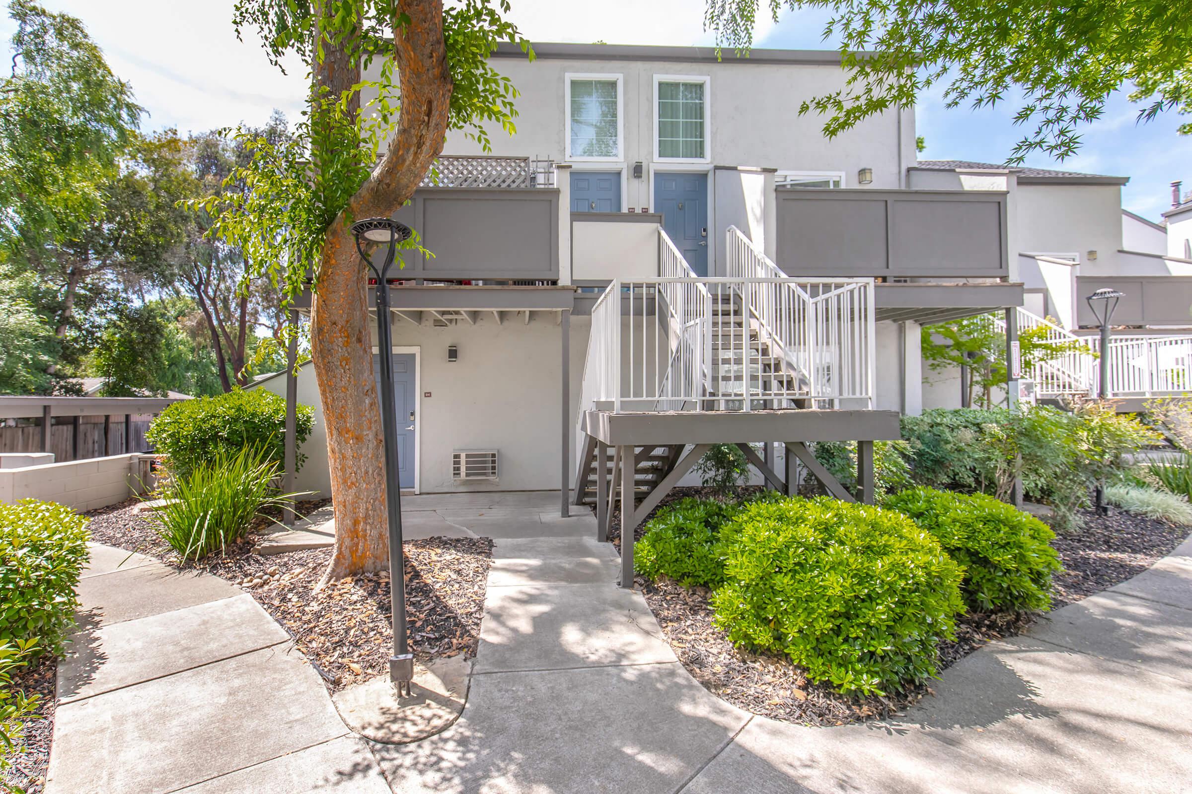 Two-story residential building with a light exterior, featuring a staircase leading to the upper level. The area is landscaped with shrubs and trees, and a pathway runs alongside. Outdoor amenities, like a lamp post and a seating area, enhance the inviting atmosphere of the property.