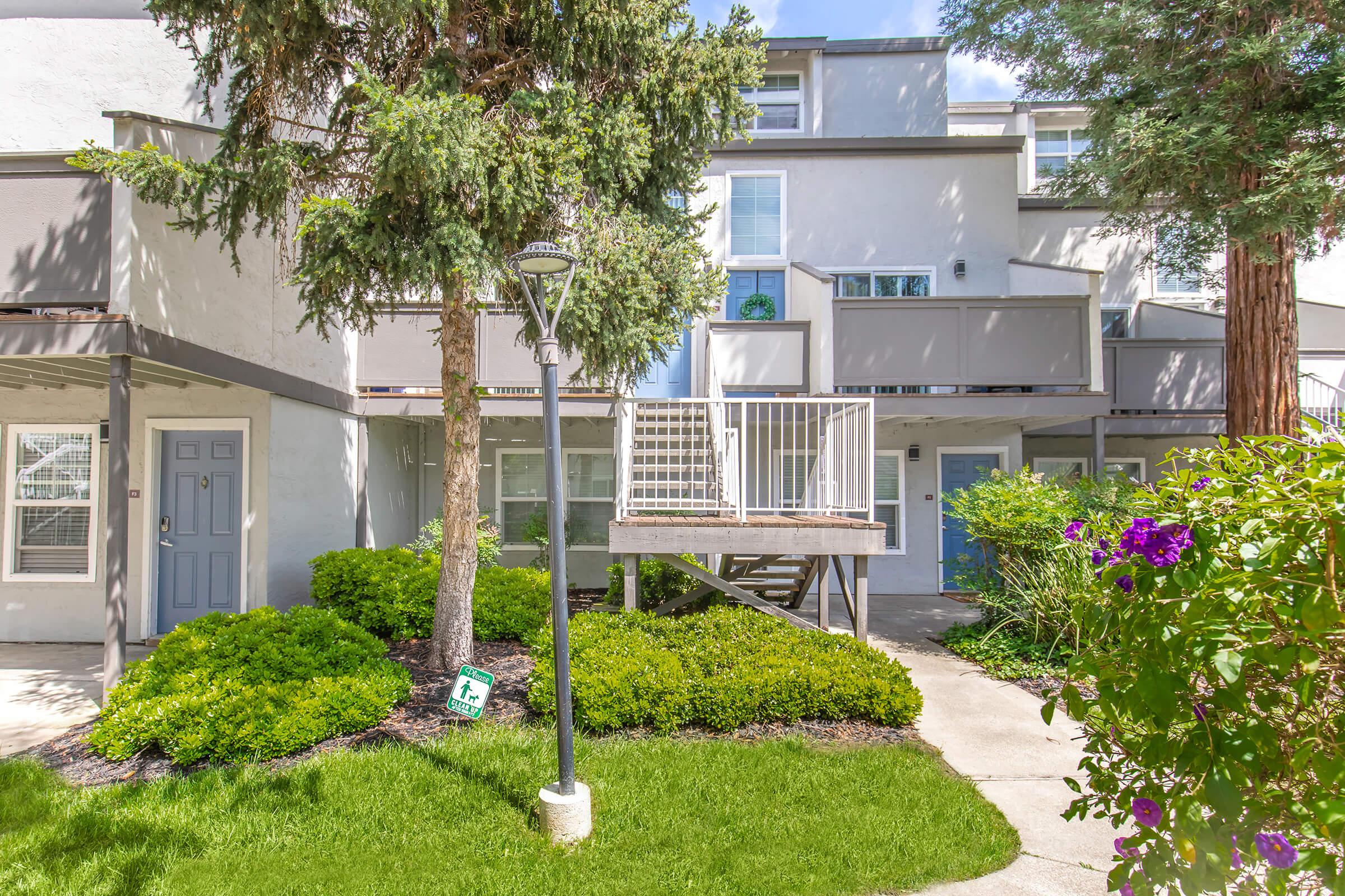 A landscaped exterior view of a multi-unit residential building featuring green grass, shrubs, and trees. The building has multiple entrances, with a wooden staircase leading to a raised deck, and a lamp post in the foreground. Bright sunlight illuminates the scene, creating a cheerful ambiance.