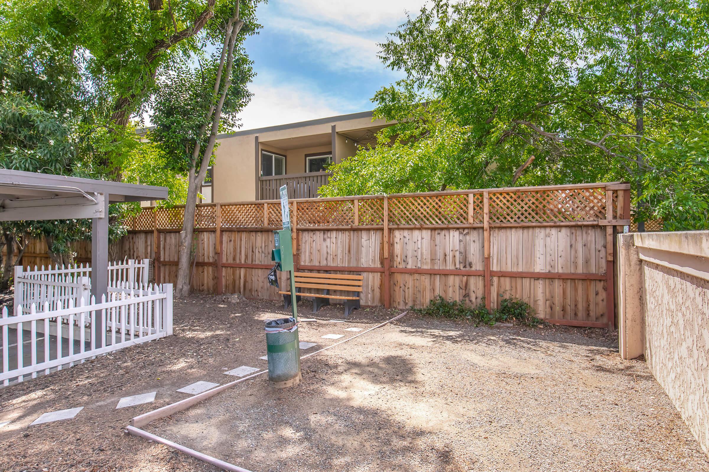 A fenced outdoor area with a gravel surface, featuring a wooden bench, a trash can, and a small section of white picket fence. Lush green trees provide shade, and a two-story building is visible in the background. The space appears clean and well-maintained, suitable for relaxation or socializing.