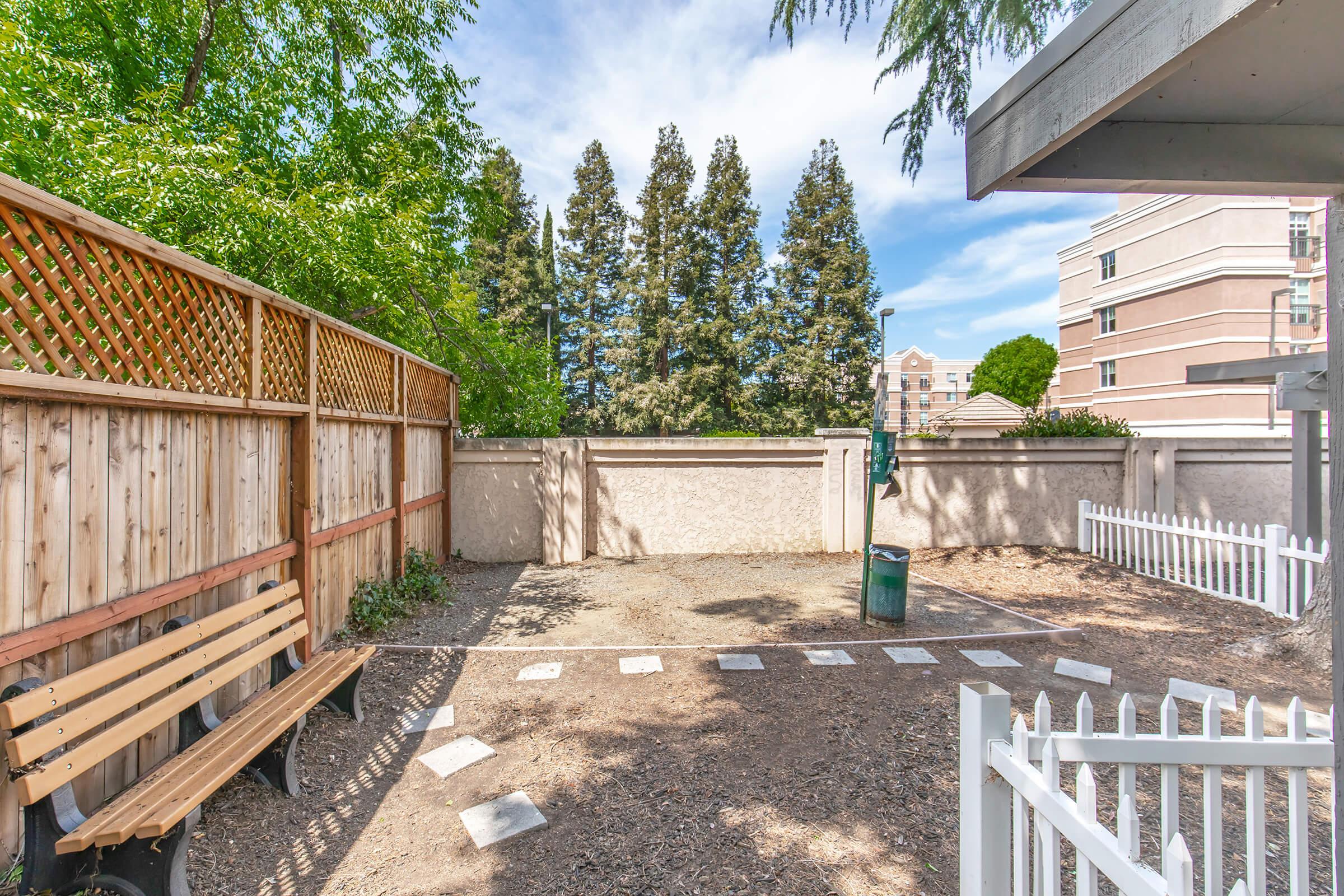 A small, enclosed yard featuring a wooden bench, gravel ground, and a green trash can. The area is surrounded by a wooden fence with lattice styling and has tall evergreen trees in the background. A low white picket fence outlines one side, and a building is visible in the distance under a partly cloudy sky.
