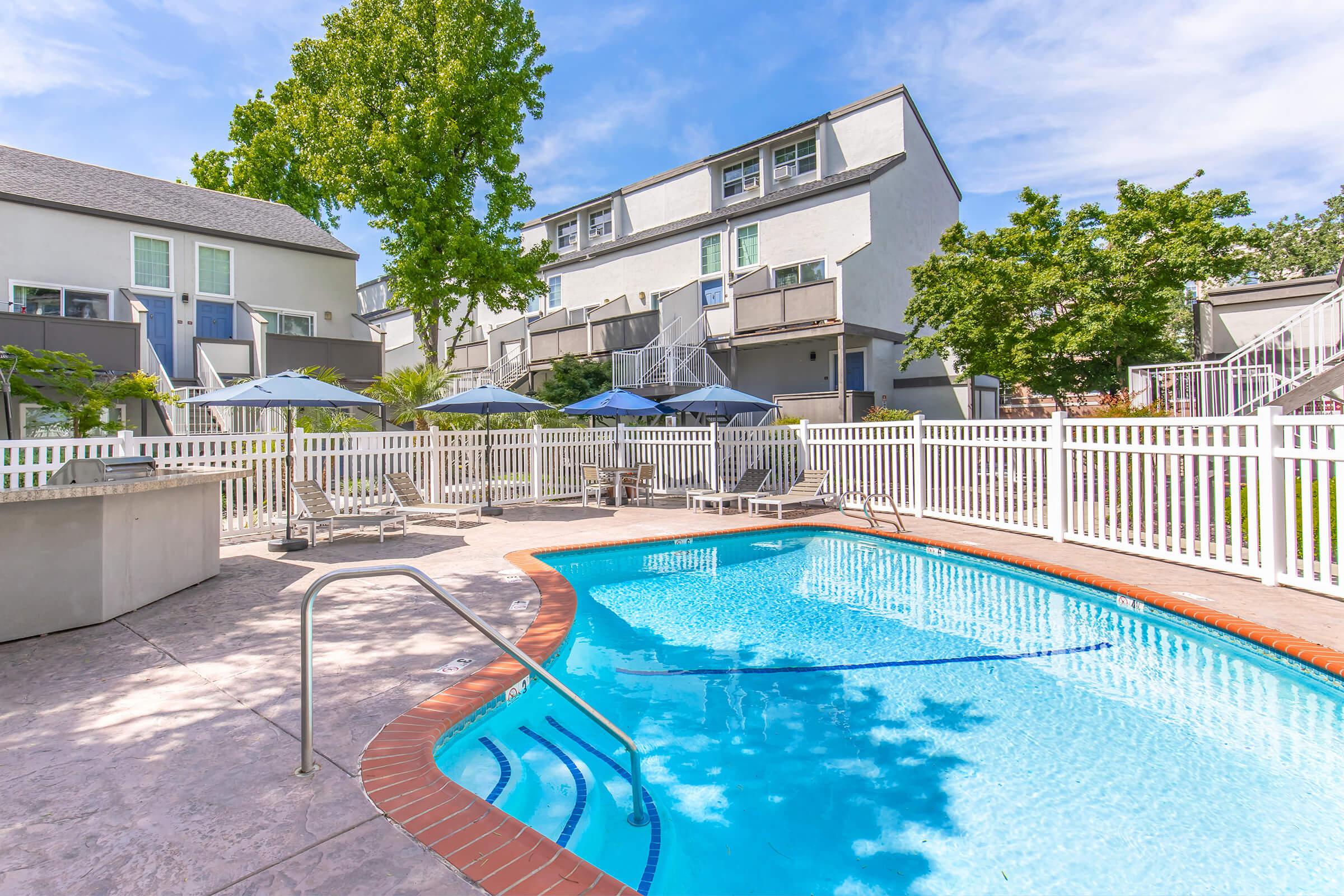 A swimming pool surrounded by a fenced area with lounge chairs and umbrellas. In the background, there are two-story apartment buildings with balconies. The sky is clear with a few clouds, and green trees are visible nearby, creating a relaxing outdoor atmosphere.