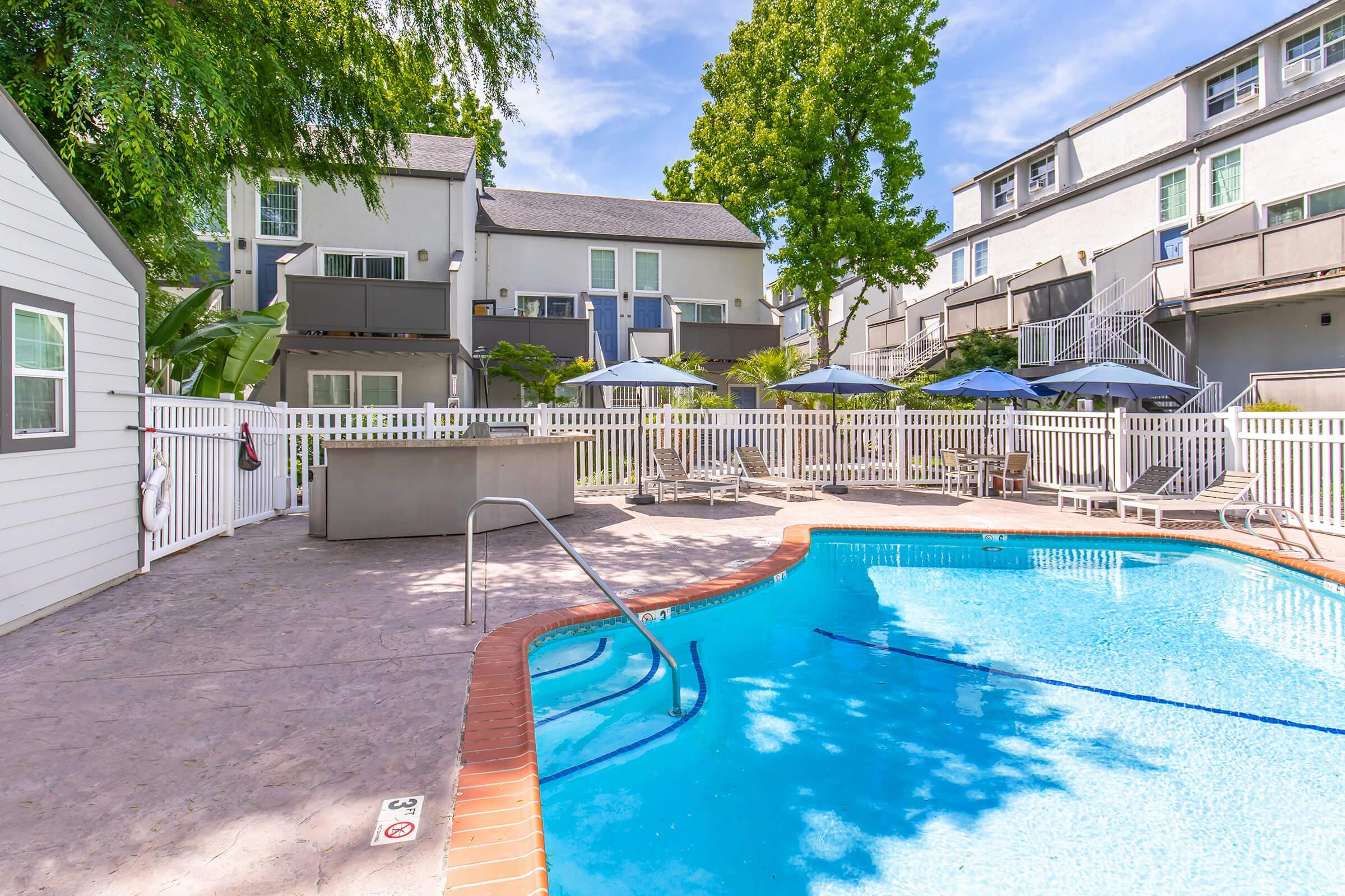 A clear blue swimming pool surrounded by a white fence, with lounge chairs and umbrellas set up. In the background, multiple two-story apartment buildings are visible, alongside lush greenery and trees under a bright blue sky.