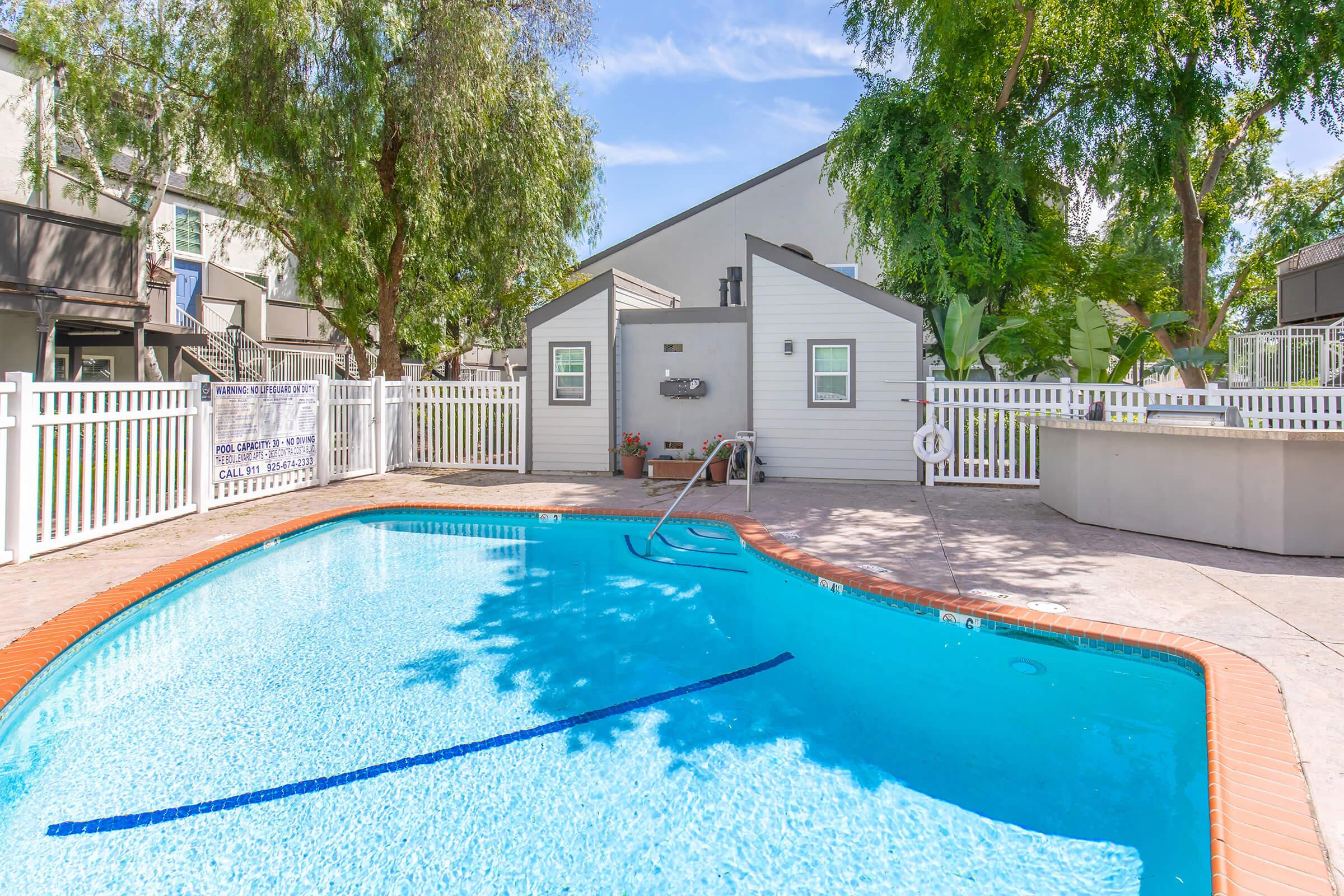 A sunny outdoor swimming pool with clear blue water, surrounded by a white fence and trees. Nearby, there are small buildings and seating areas. The pool area features a ladder for entry and lush greenery, creating a relaxing atmosphere.