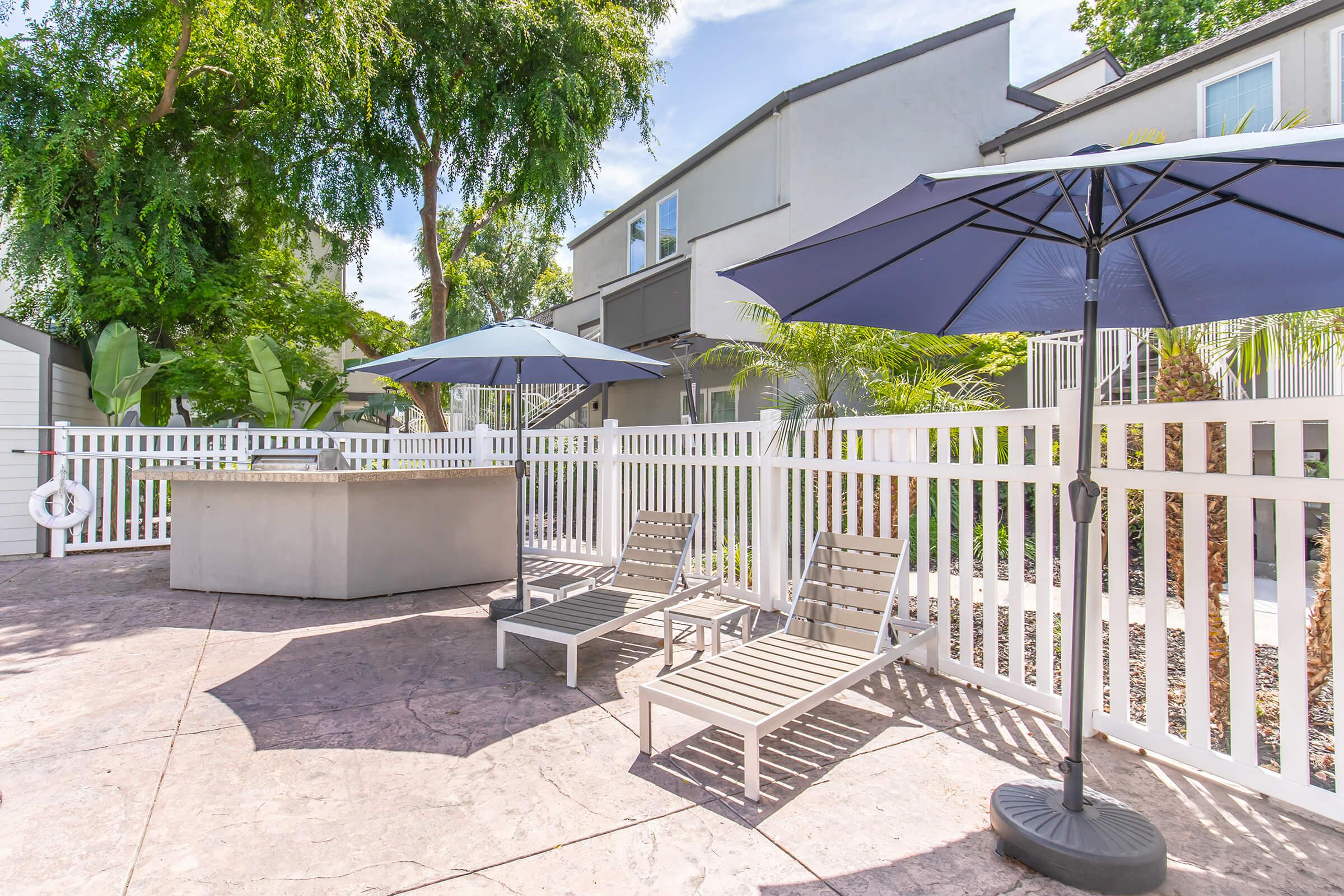 A tranquil outdoor space featuring two lounge chairs beneath blue umbrellas, a modern hot tub, and a white picket fence. Lush greenery and a stylish building are visible in the background, creating a relaxing atmosphere perfect for leisure and enjoyment.