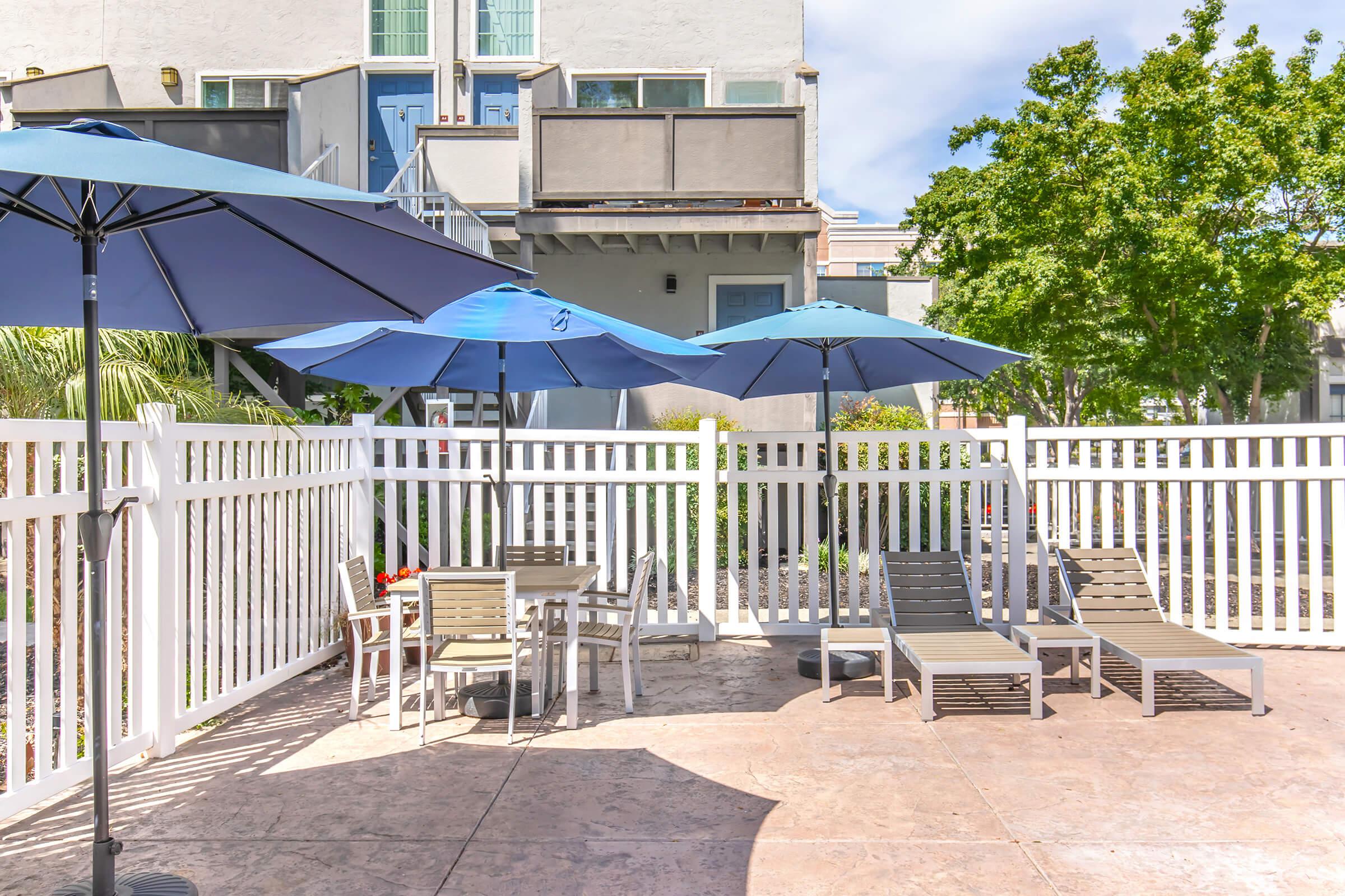 A poolside area featuring lounge chairs and blue umbrellas, surrounded by a white picket fence. In the background, a building with windows and doors is visible, along with green trees and landscaping. The scene is bright and inviting, perfect for relaxation.