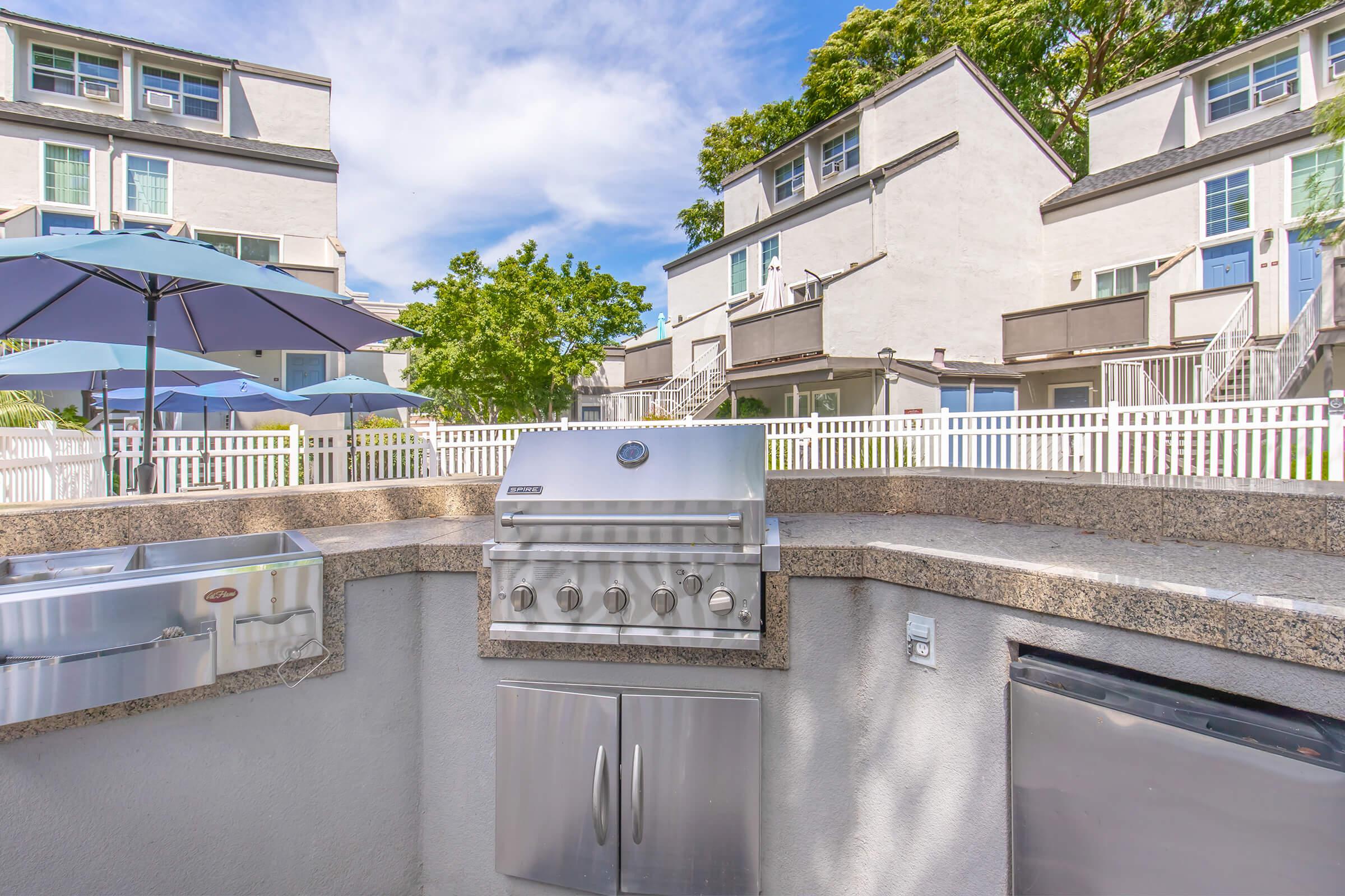 Outdoor grilling area featuring a stainless steel barbecue grill, a food prep station, and a large umbrella for shade. In the background, there are multiple modern townhouses with white exteriors and a well-maintained lawn, under a clear blue sky.