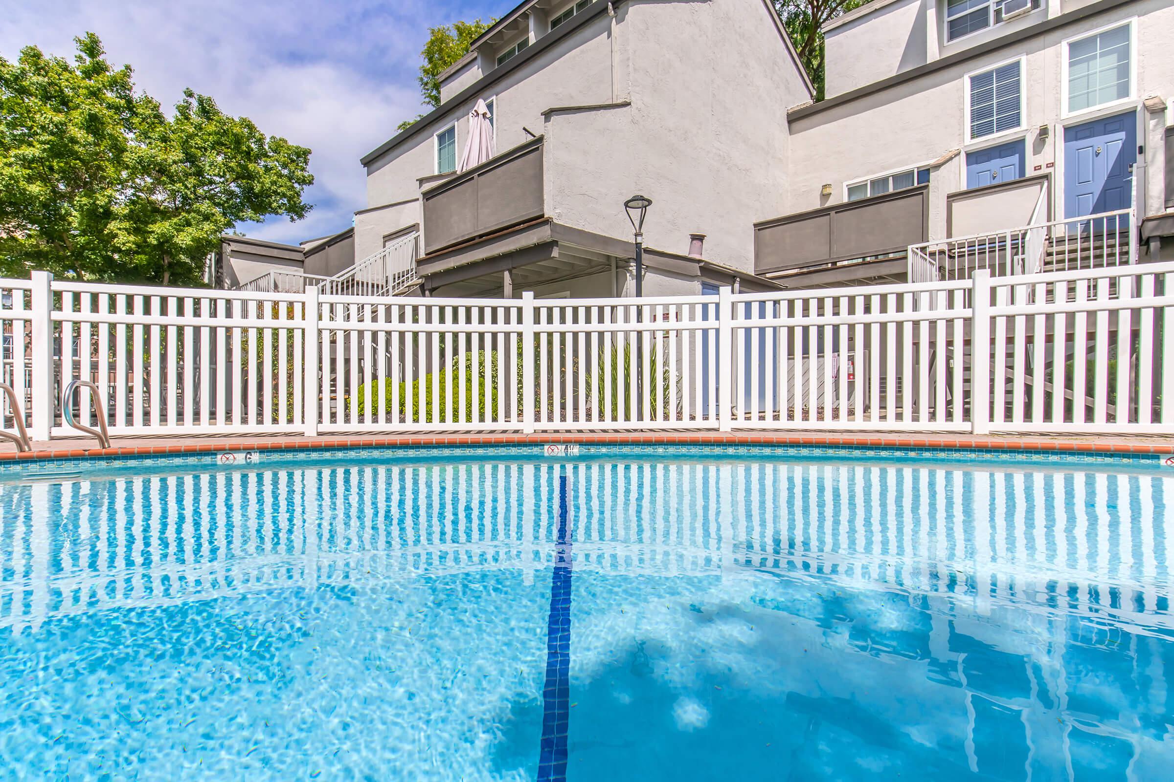 A clear swimming pool surrounded by a white picket fence, with two buildings visible in the background. The scene is bright with blue skies and greenery nearby, creating a relaxing atmosphere.