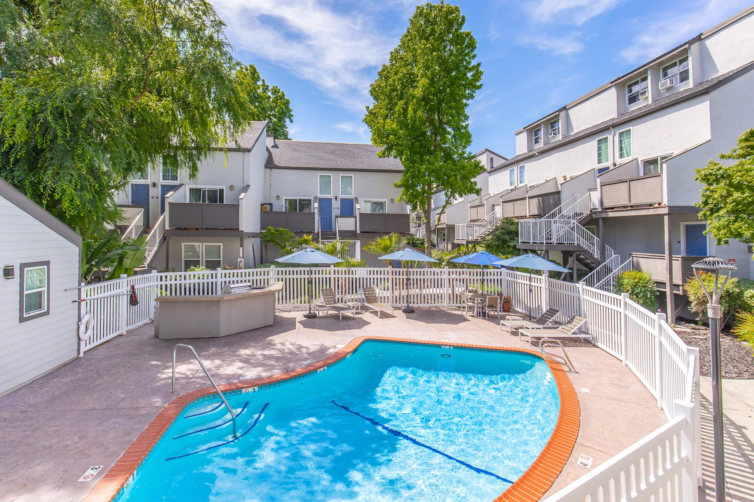 A sunny courtyard featuring a blue swimming pool surrounded by lounge chairs. White fencing encloses the pool area, with several umbrellas providing shade. In the background, there are multi-story buildings with balconies and lush green trees. Clear blue skies complete the tranquil scene.