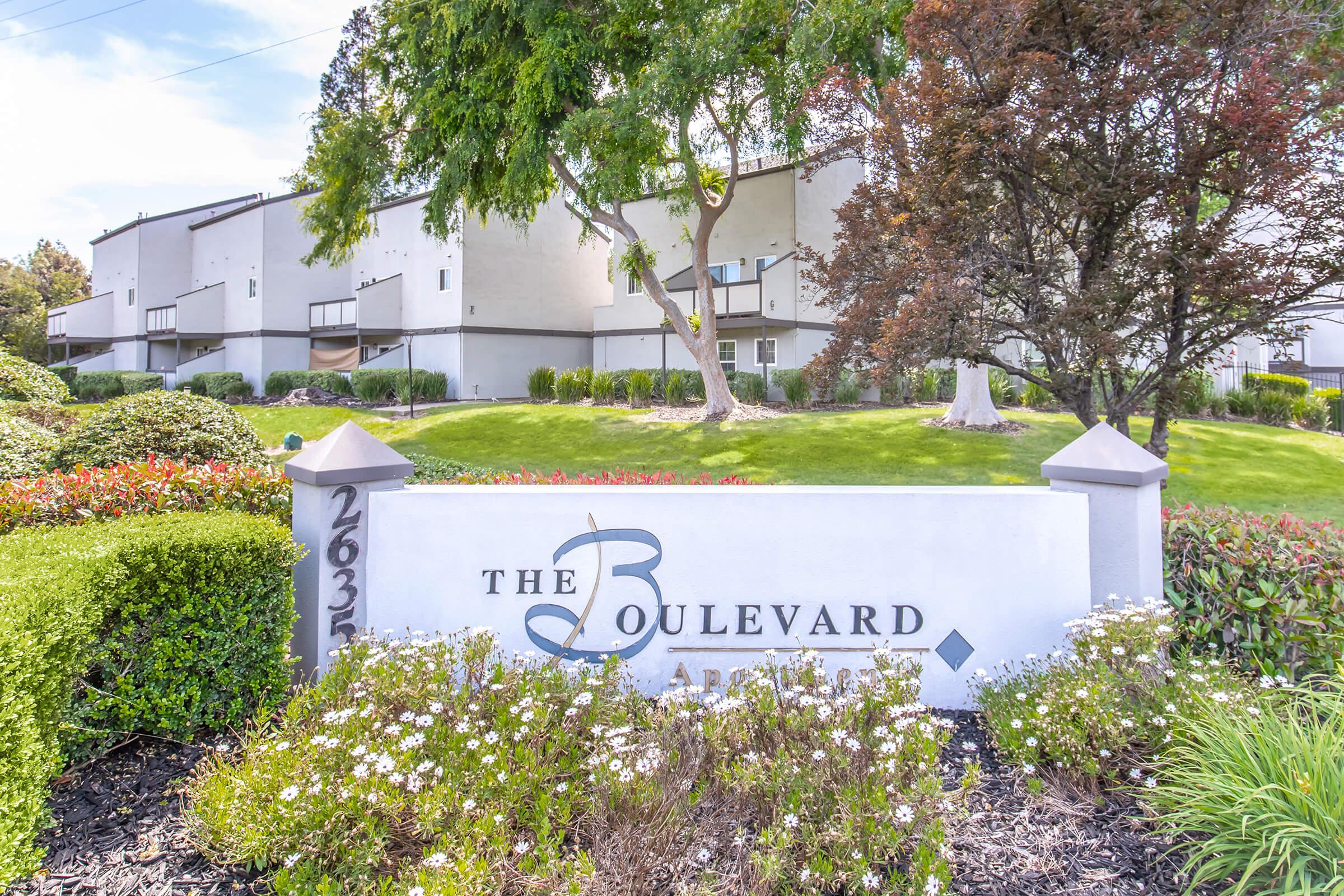 Sign for "The Boulevard Apartments" with the address 2635, surrounded by well-maintained landscaping including bushes and flowers, and a backdrop of apartment buildings and trees. The setting appears serene and inviting, highlighting the complex's welcoming atmosphere.