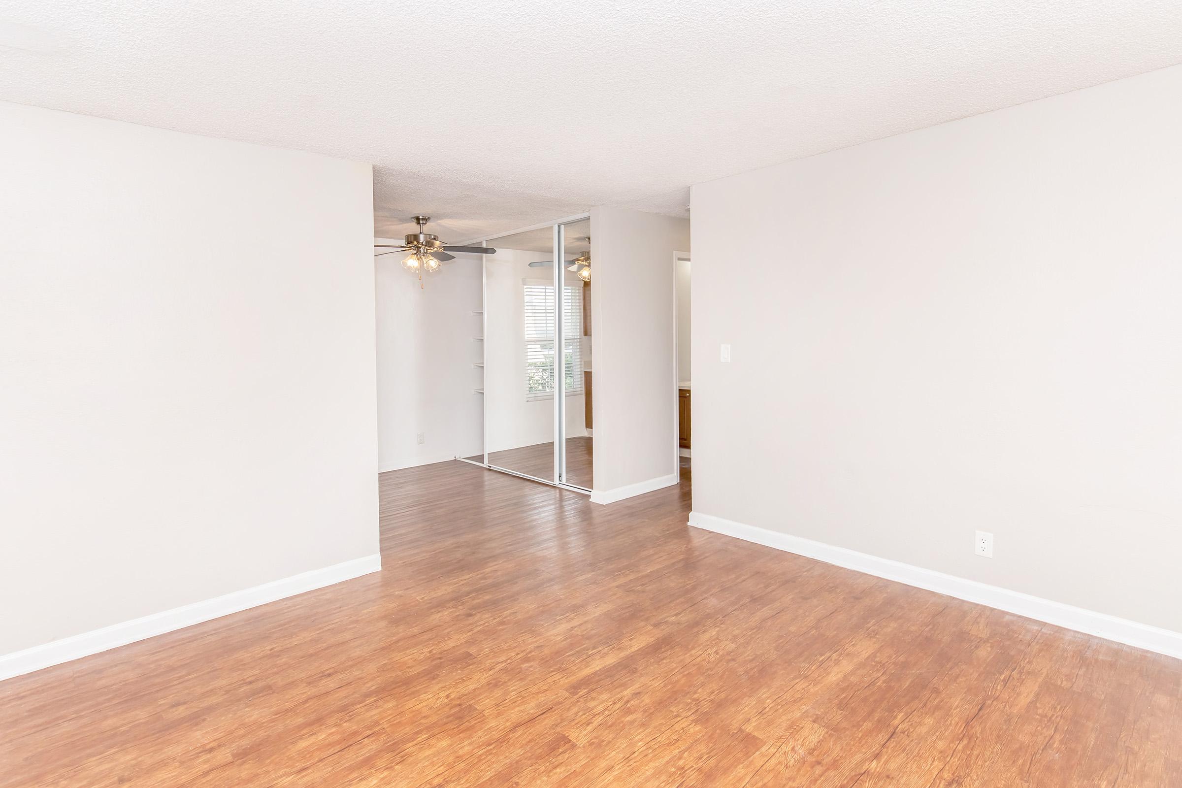 A spacious, empty room with light-colored walls and a wood-like floor. The area features a ceiling fan and a mirror closet on one side. Natural light filters in through a nearby window, enhancing the bright and airy atmosphere of the space.