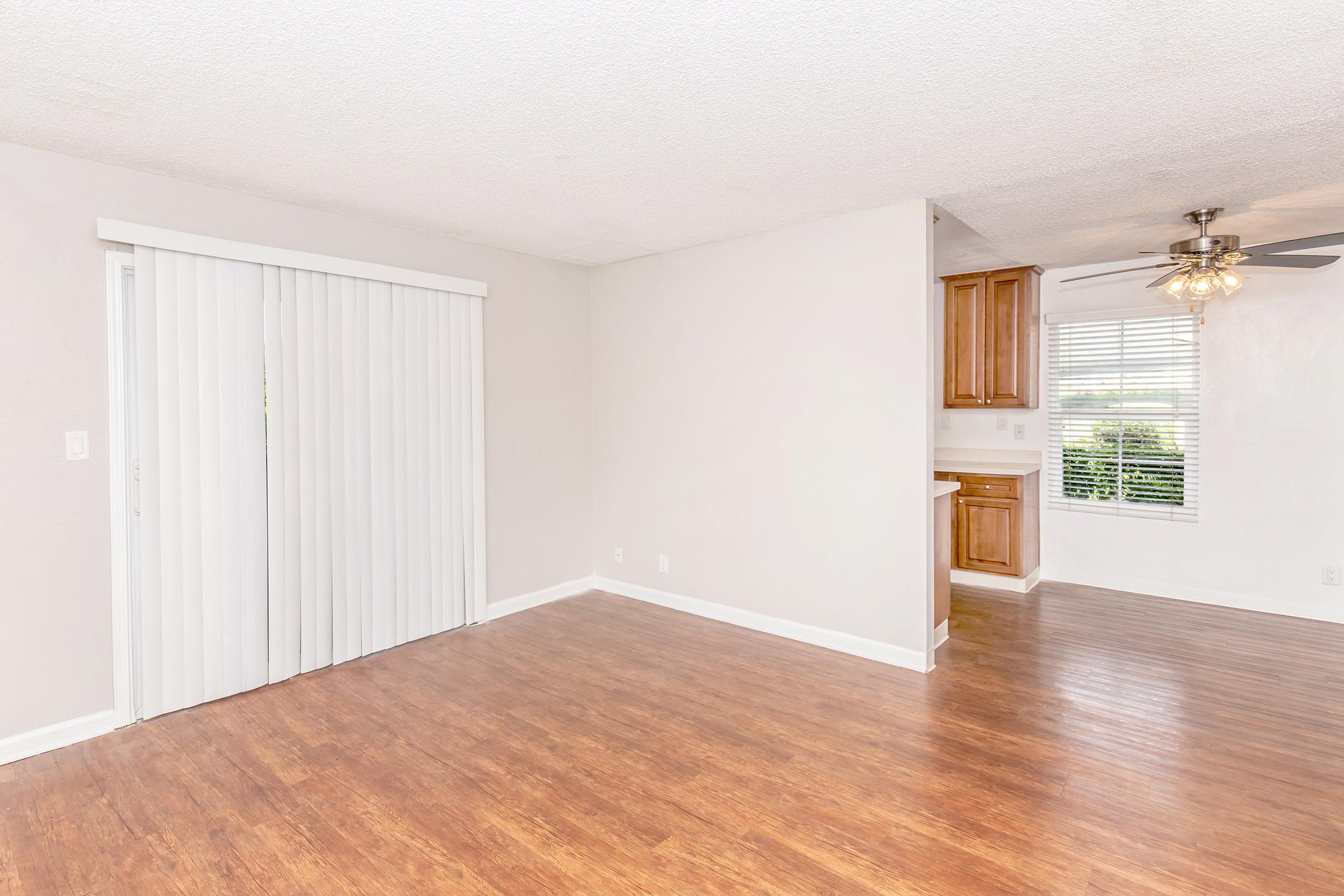 Interior of a light-colored living room featuring laminate wood flooring, a sliding glass door with vertical blinds, and an open doorway leading to a kitchen area with wooden cabinetry. The walls are painted in a soft, neutral tone, and a ceiling fan is visible. Natural light filters in through the window.