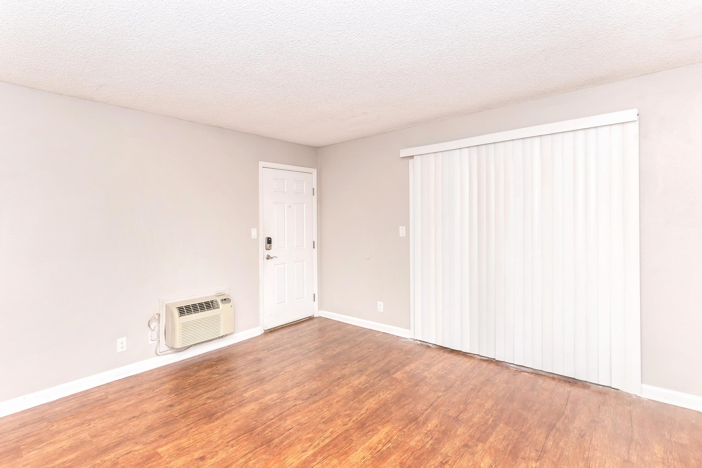 A spacious and empty room with light gray walls and wooden flooring. It features a wall-mounted air conditioning unit, a white door leading outside, and a large window covered with vertical blinds. The overall atmosphere is bright and uncluttered, ideal for various uses.