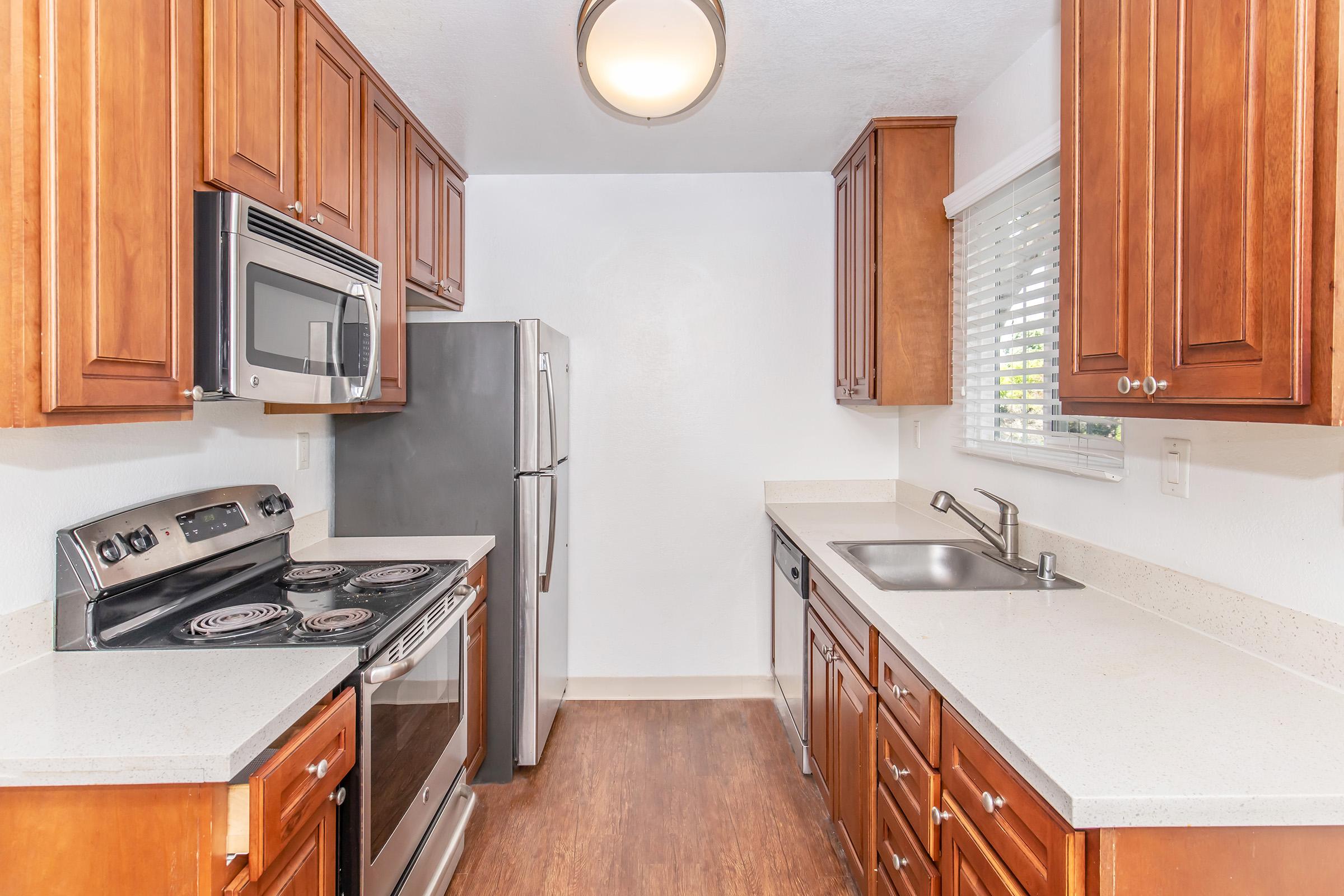 Modern kitchen featuring wooden cabinets, stainless steel appliances including a microwave and refrigerator, a silver stove with an oven, and a sink. The countertop is light-colored, with a window adorned with blinds. The flooring is a warm, wooden texture, creating a bright and inviting atmosphere.