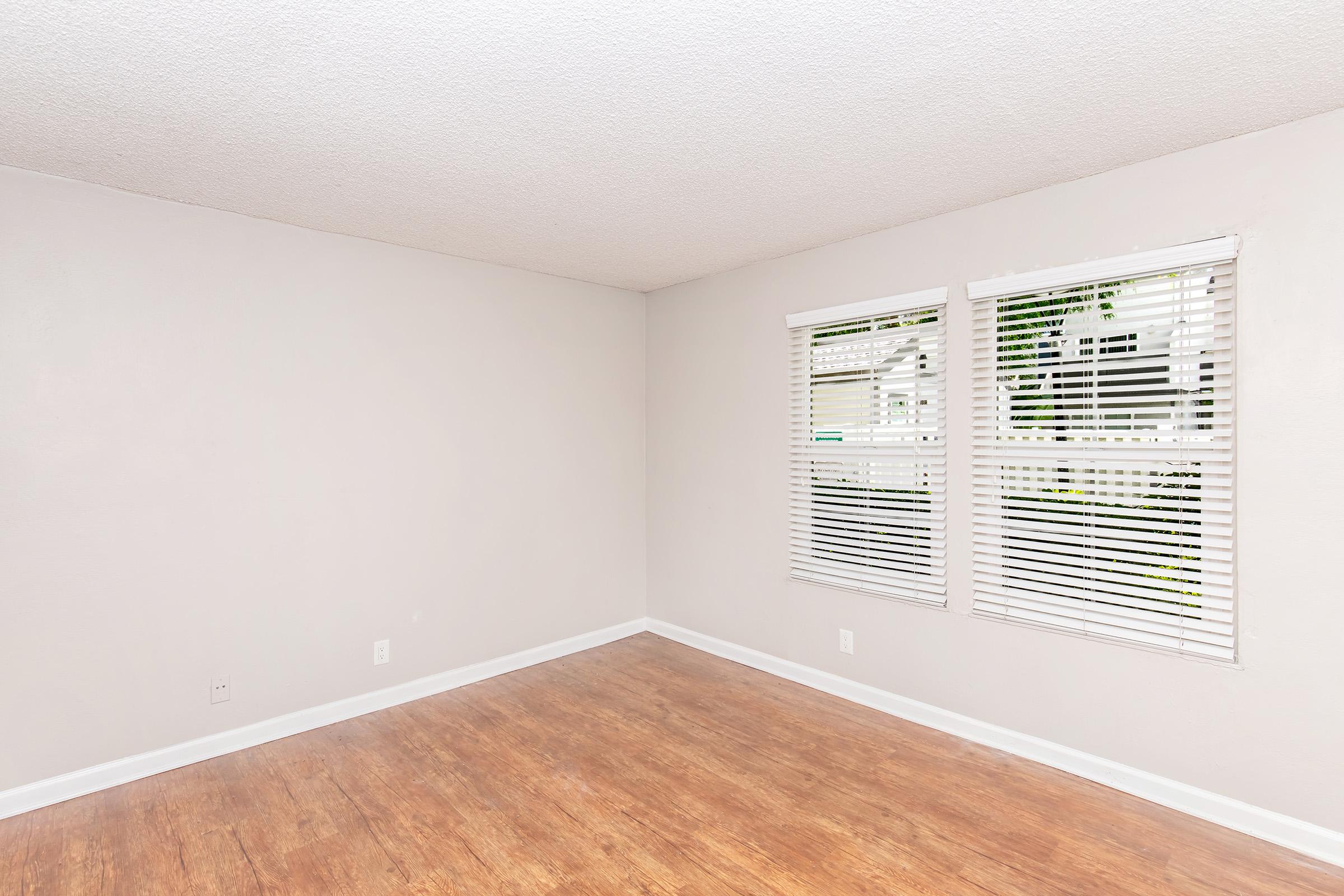 Empty room with light-colored walls and wooden floor. Two windows with white blinds allow natural light to enter, creating a bright atmosphere. The space appears clean and ready for furniture placement.