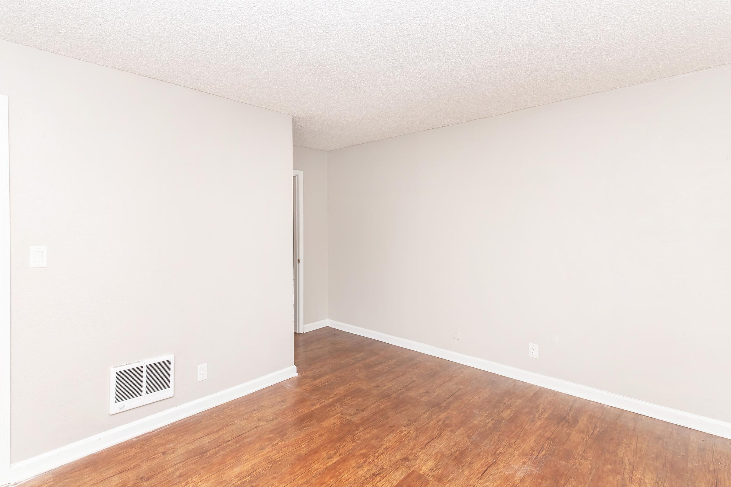 An empty room featuring light gray walls and a smooth wood floor. There is a vent on the wall, and a door leading to another space is visible in the background. The room is well-lit and lacks furniture or decorations, providing a minimalistic and open feel.
