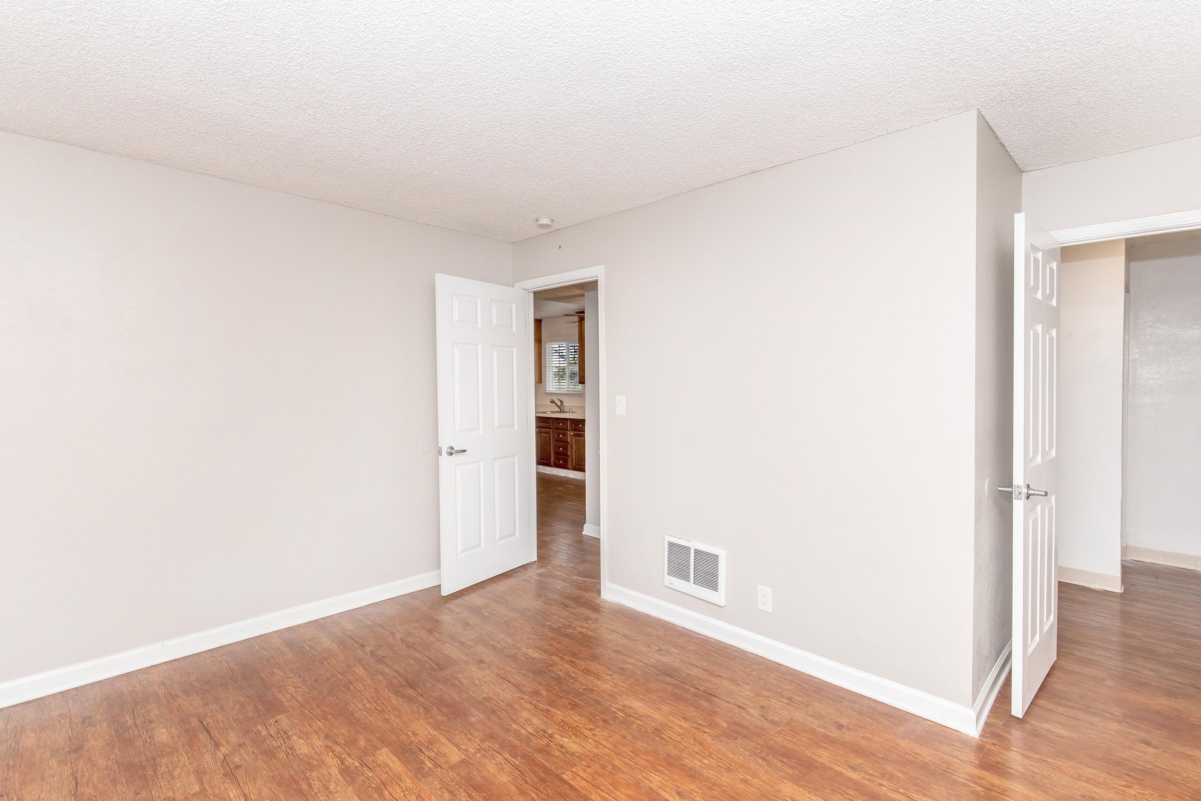 A view of a small, empty room with light-colored walls and wooden flooring. There are two open doors leading to adjacent areas, one displaying a kitchen space with cabinetry. The room has a neutral, inviting aesthetic, suitable for various purposes such as a living area or office. Natural light enhances the ambiance.