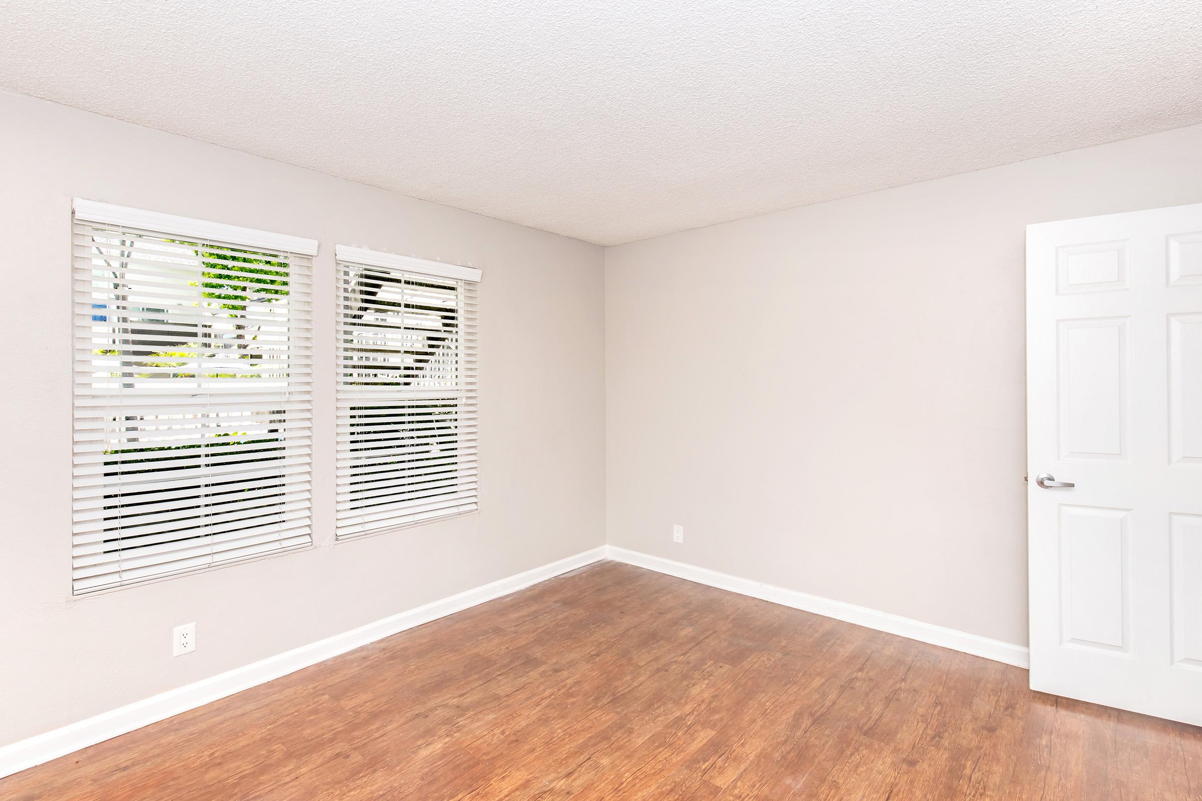 A well-lit, empty room with light beige walls and a brown wooden floor. Two window blinds allow natural light in, and a white door is visible on the right side. The space is free of furniture, creating an open and inviting atmosphere.