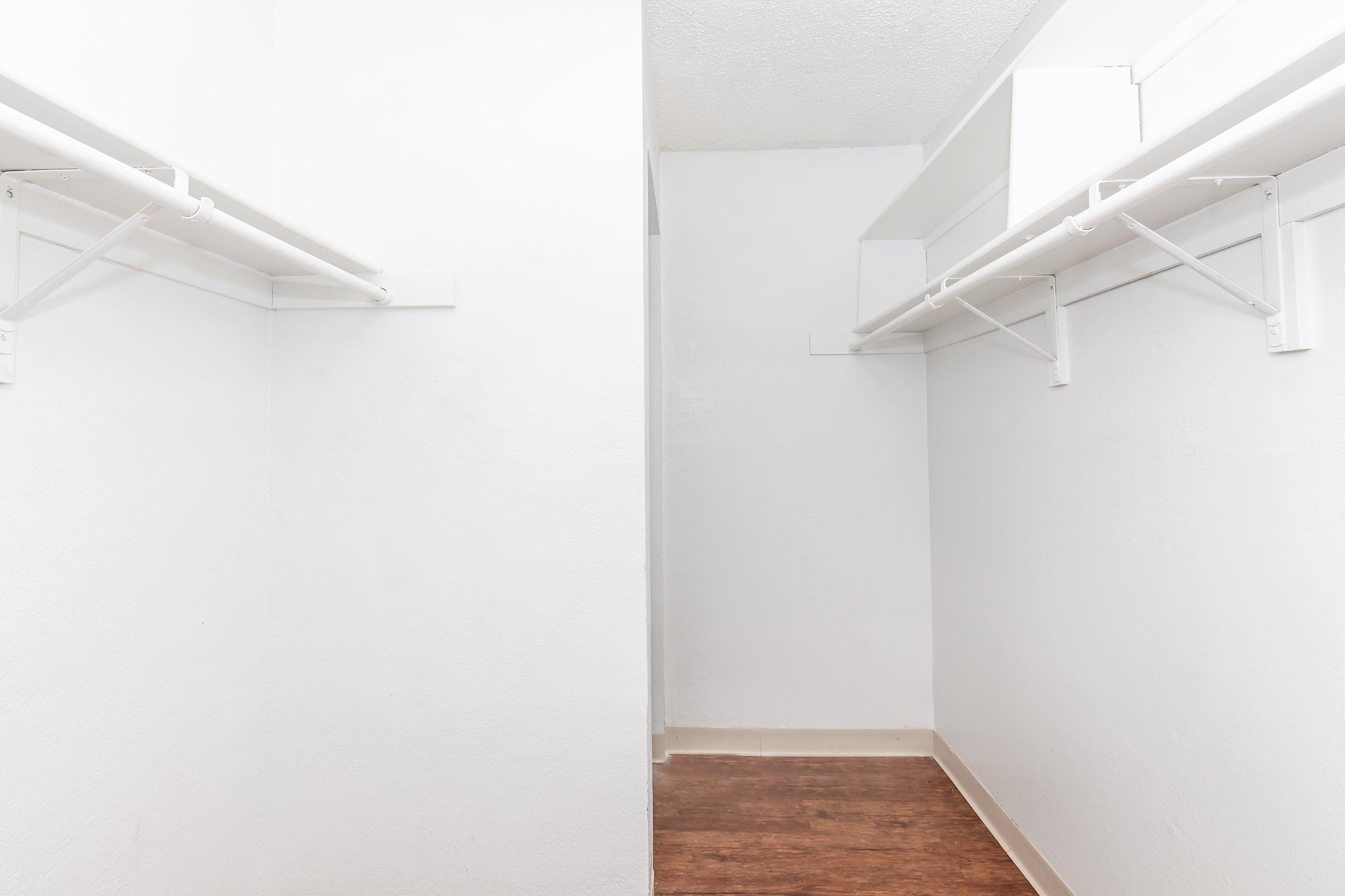 Empty interior of a closet with white walls and a wooden floor. Two sides feature hanging rods for clothes storage, while the space between them appears open and uncluttered.
