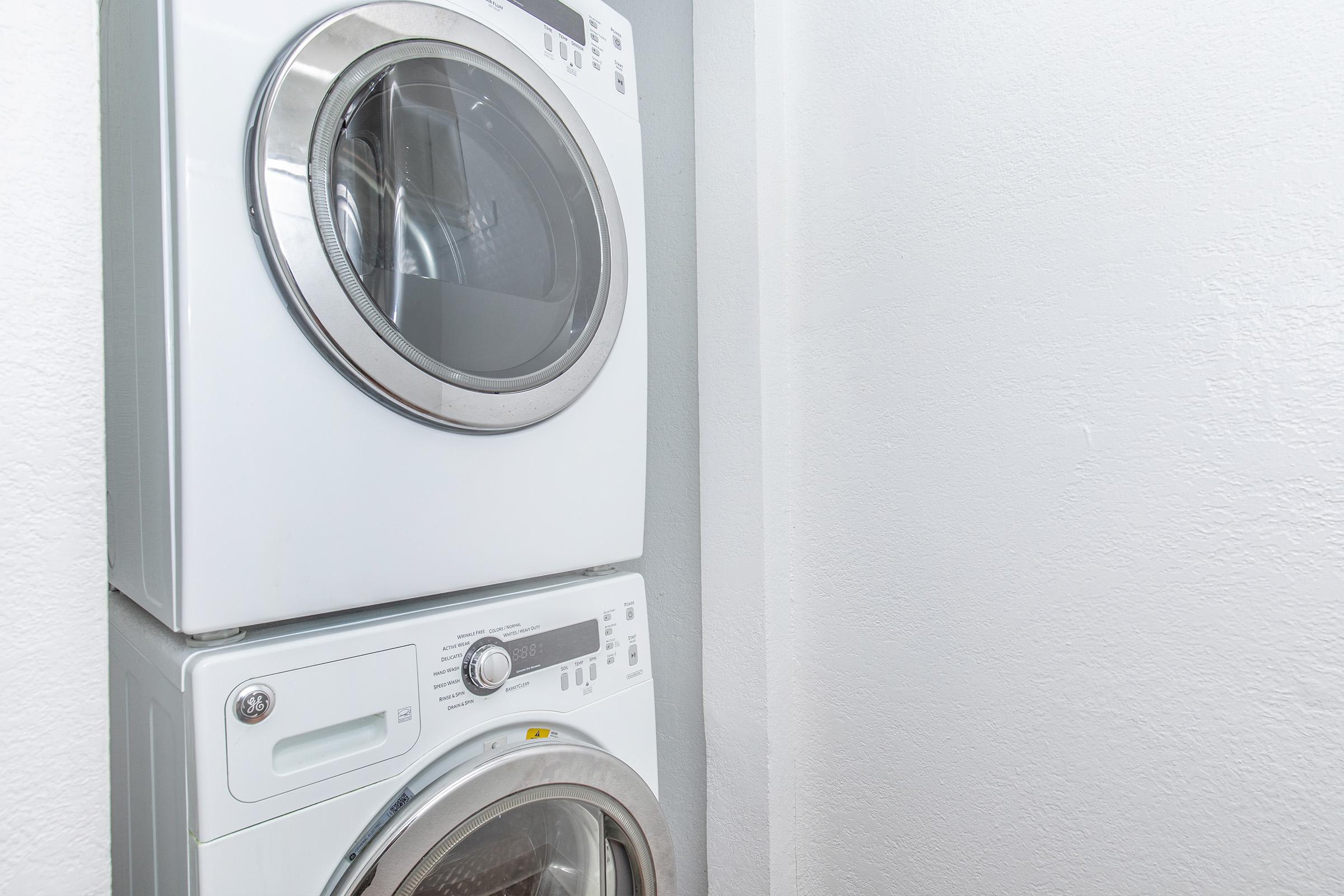 A stacked washer and dryer unit in a small laundry space. The white appliances are modern and sleek, positioned against a light-colored wall. The dryer is on top, featuring a round glass door, while the washer below has a front-loading design with various control buttons and a digital display.