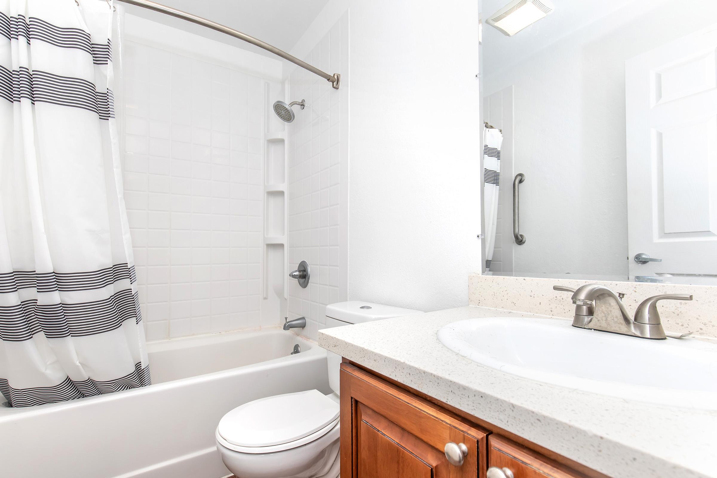 A clean bathroom featuring a white bathtub with a shower curtain, a modern sink with a wooden cabinet underneath, and a large mirror. The decor is minimalistic, with white tiled walls and light fixtures. The overall appearance is bright and well-maintained.