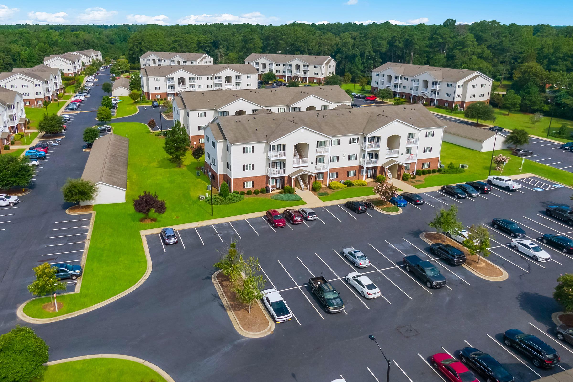 Aerial view of a residential apartment complex surrounded by green lawns and trees. There are multiple buildings with balconies and parking lots filled with cars. The area features well-maintained landscaping and clear blue skies in the background.