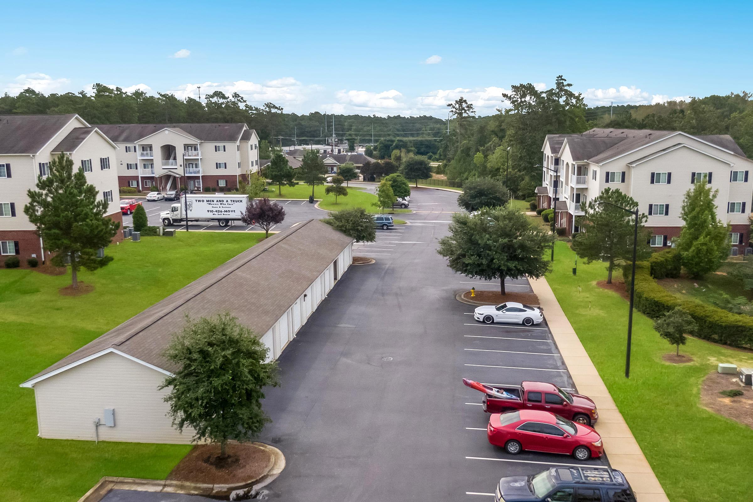 Aerial view of a residential area featuring two-story apartment buildings, well-maintained green lawns, and a parking lot with several cars. Trees are planted along the pathways, and a clear blue sky is visible in the background.