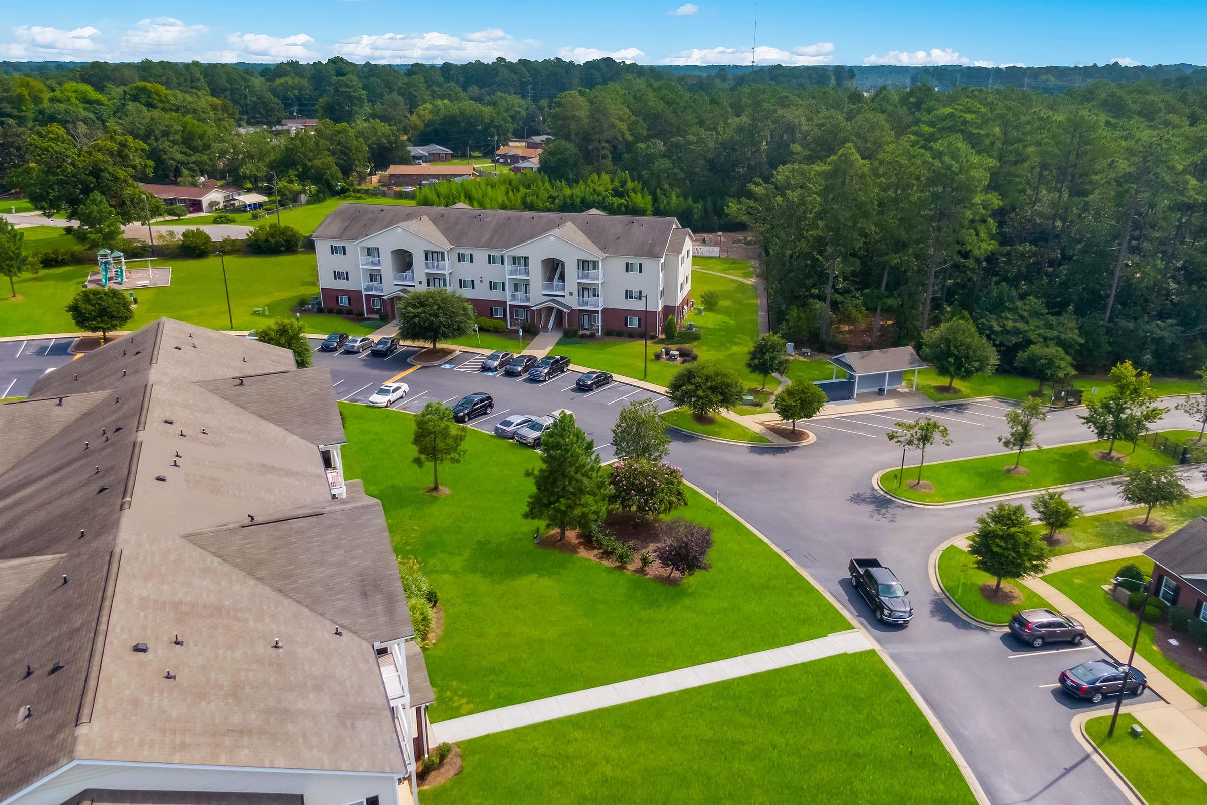 Aerial view of a residential area featuring landscaped lawns, multiple apartment buildings, and a parking lot filled with cars. Surrounding greenery includes trees and open spaces. The sky is clear with some clouds, suggesting a pleasant day.