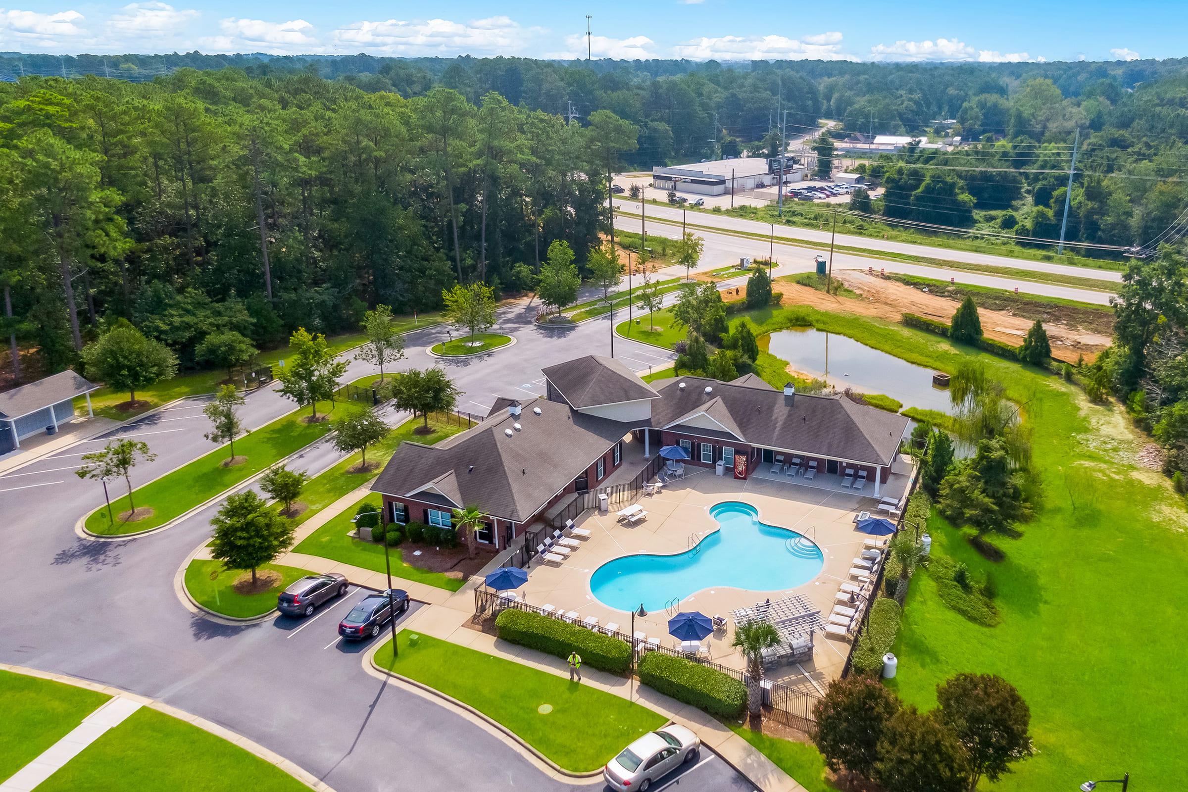 Aerial view of a residential complex featuring a swimming pool surrounded by lounge chairs. The building has a modern design with green lawns and trees. Nearby, there are parked cars and a road visible, along with a pond and undeveloped land in the background.