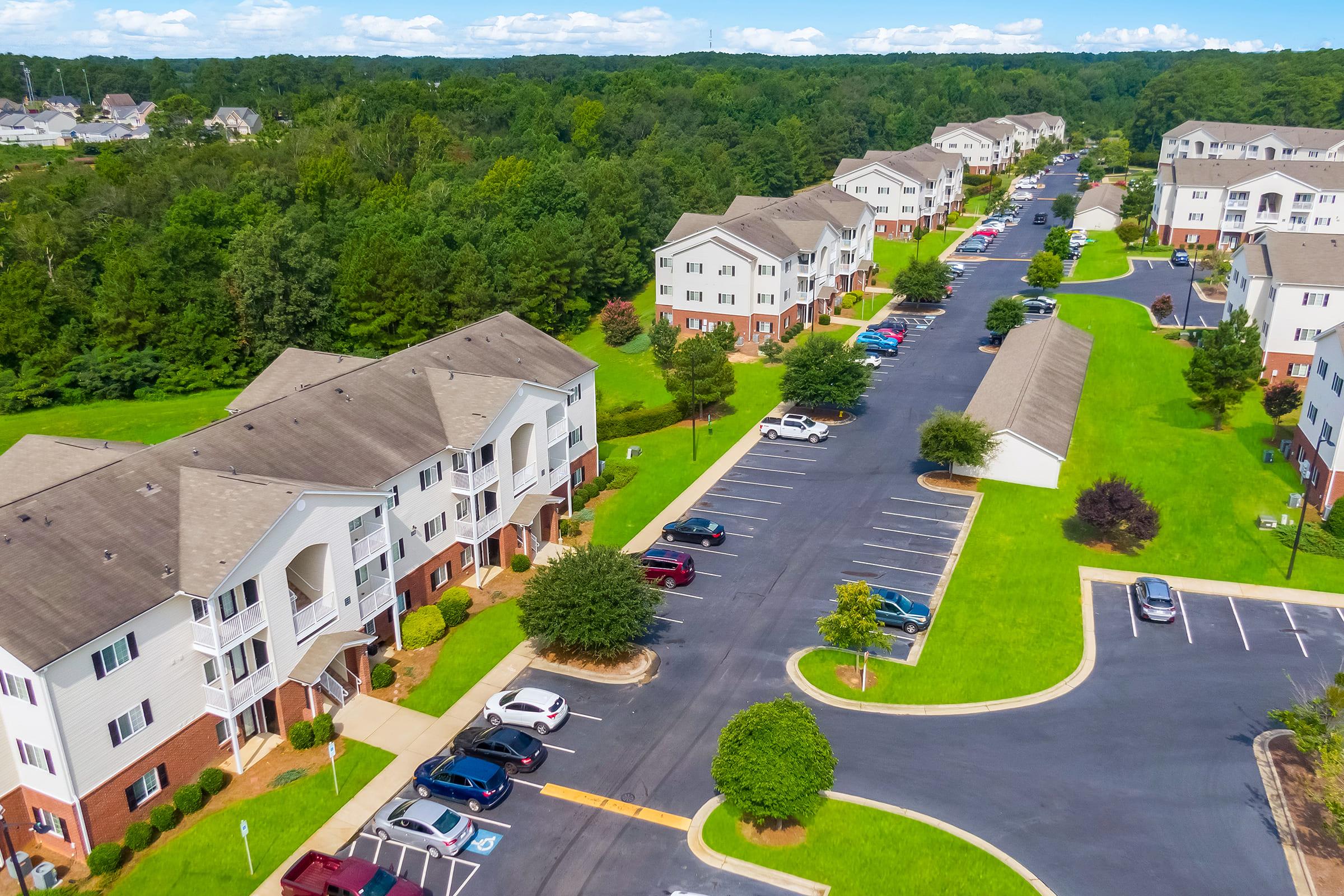 Aerial view of a residential area with modern apartment buildings surrounded by green lawns and trees. The image shows a well-maintained parking lot with several parked cars along a paved road, suggesting a suburban neighborhood with ample outdoor space and landscaping.