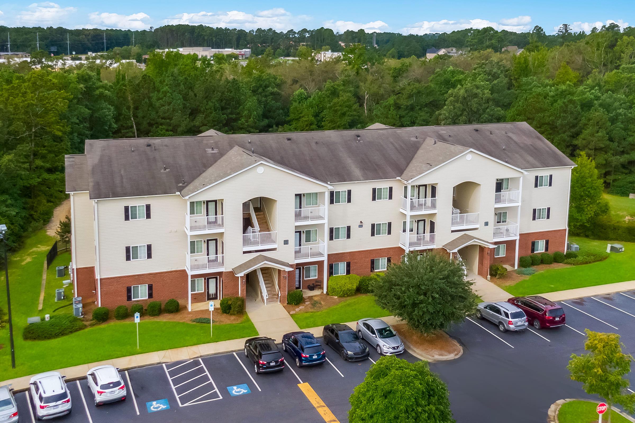 Aerial view of a multi-unit apartment building surrounded by greenery. The building has a mix of balconies and large windows, with a parking area in front that contains several cars. The scene is set against a backdrop of trees and a clear blue sky, indicating a suburban neighborhood.