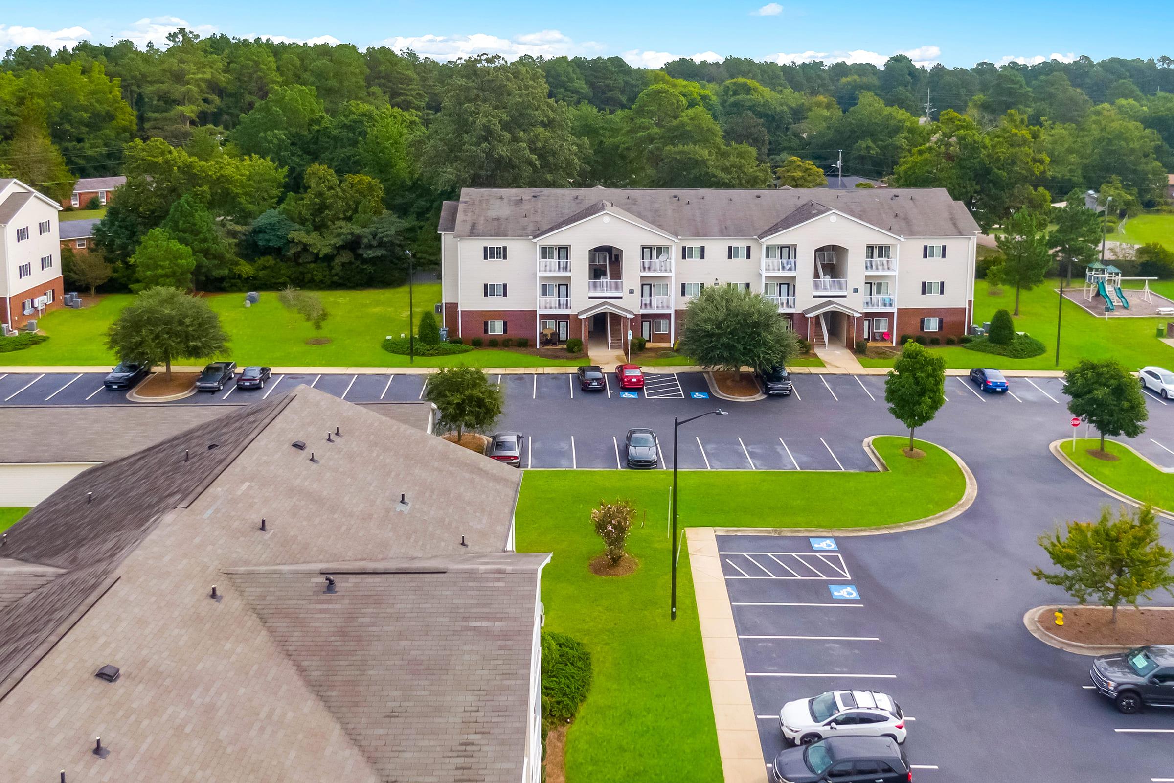 Aerial view of a residential apartment complex surrounded by green trees. The image shows multiple buildings with parking spaces, including designated handicapped spots. There are cars parked in several spaces, and a playground is visible in the background, adding to the community setting.