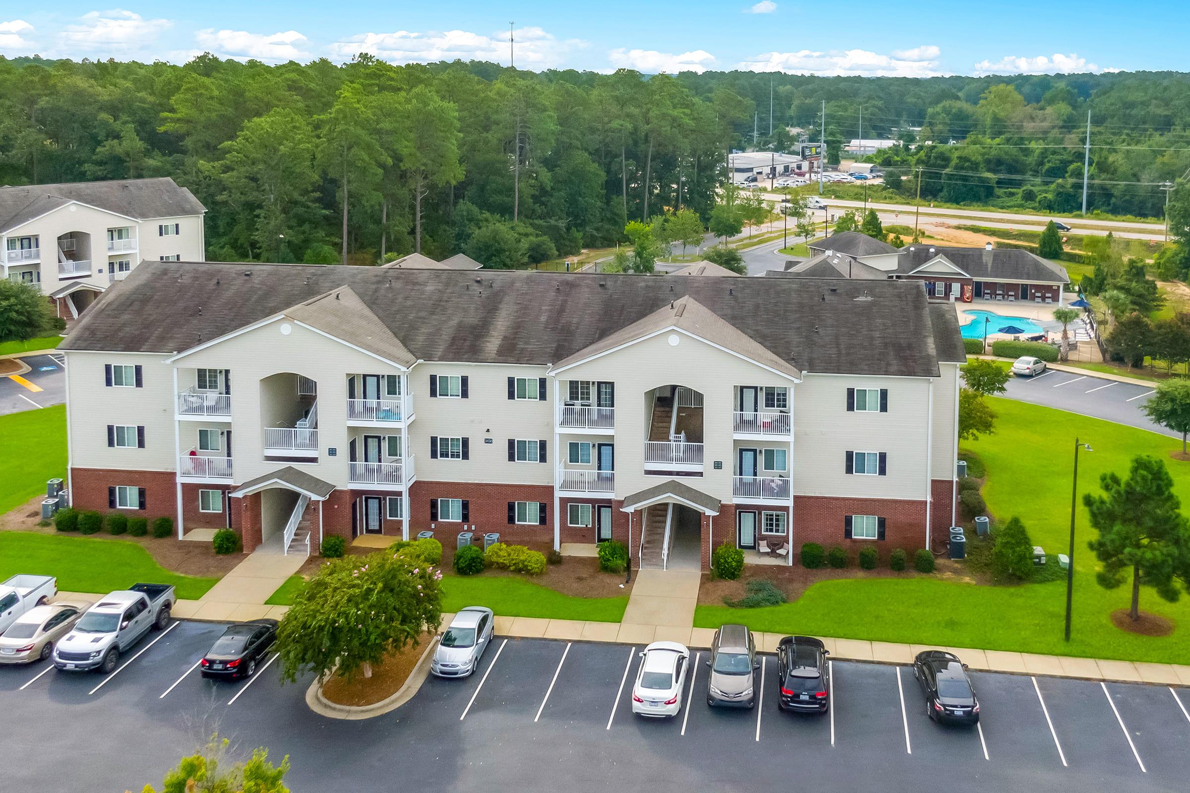 Aerial view of a multi-unit apartment building surrounded by green trees, with a parking lot in the foreground filled with parked cars. The building features a mixture of brick and siding exteriors, and there is a swimming pool visible in the background, along with additional residential structures.