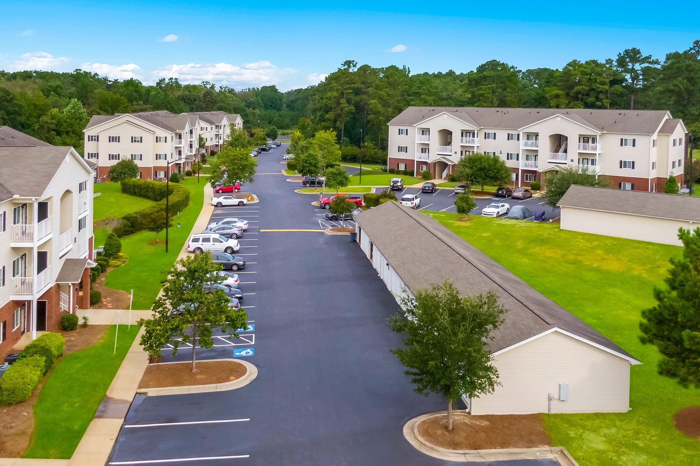Aerial view of a residential complex featuring several multi-story apartment buildings surrounded by green grass and trees. A paved parking lot with parked cars lines the street, and pathways lead between buildings. Clear, blue sky overhead, creating a serene suburban atmosphere.