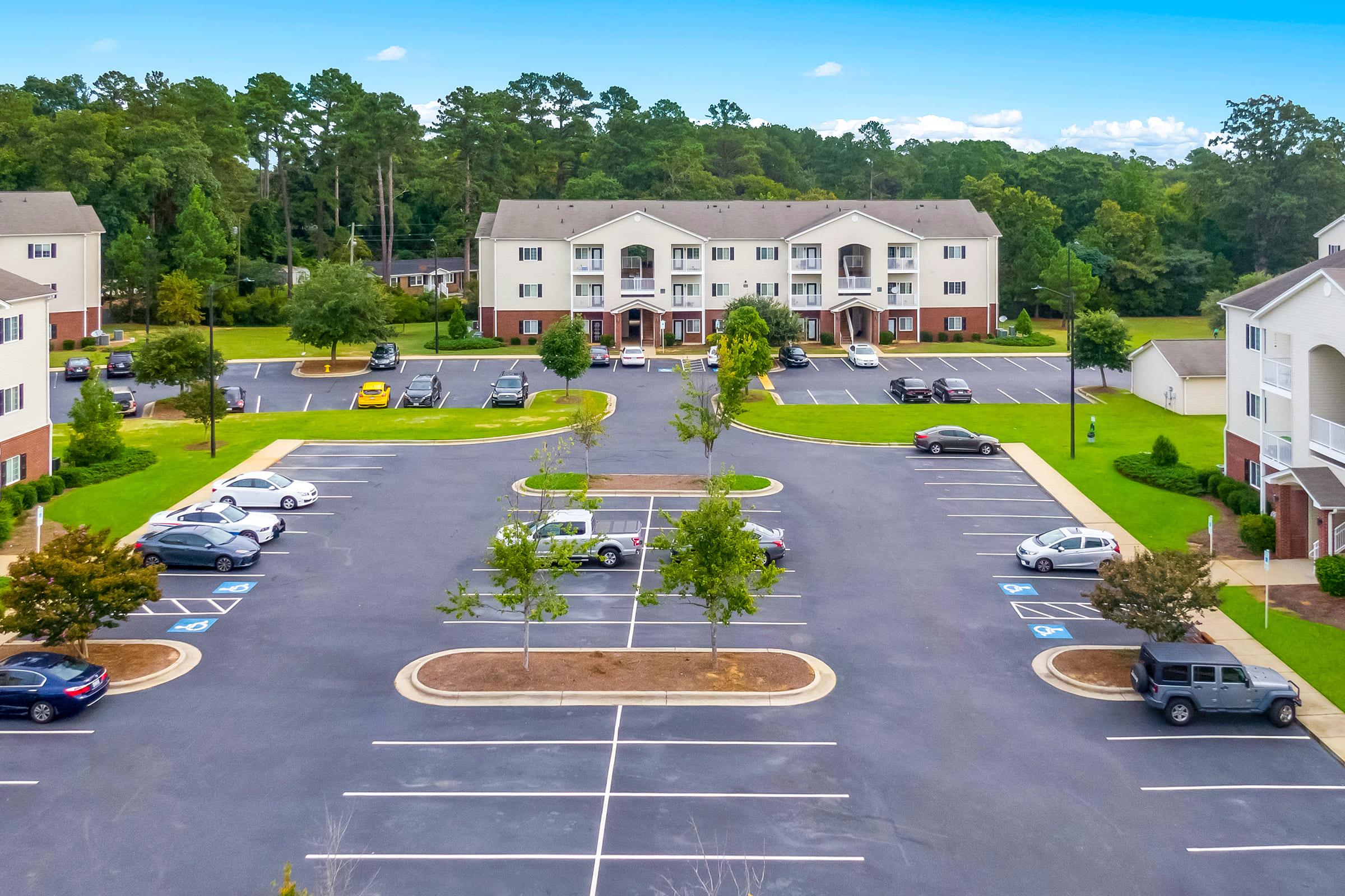 Aerial view of a residential complex featuring multiple two-story buildings surrounded by green lawns and trees. The parking lot is located in the foreground, showing several parked cars, with designated spaces visible. The scene highlights a clear blue sky and a peaceful suburban setting.