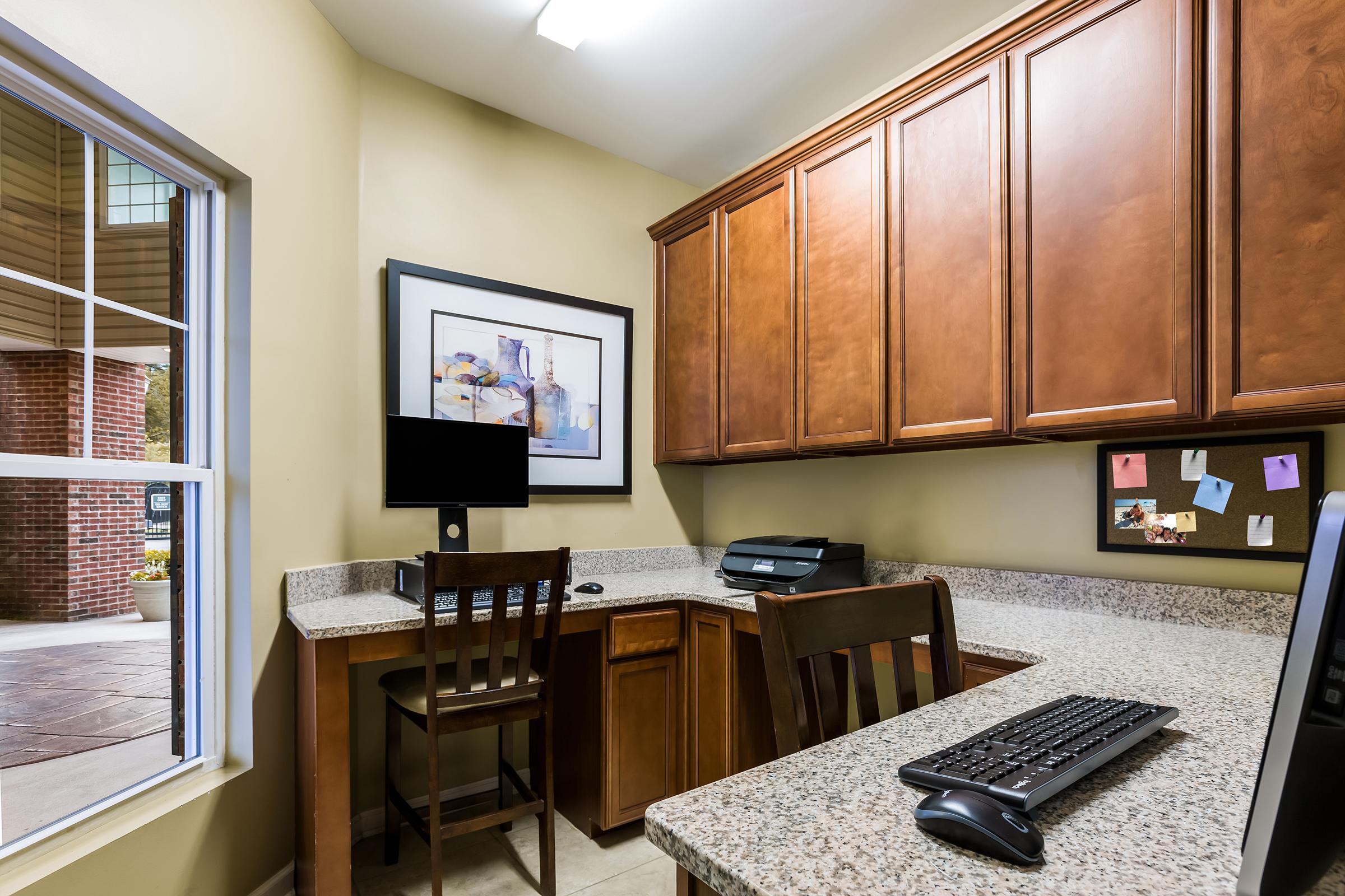 A cozy workspace featuring a desktop computer, printer, and a wooden chair against a granite countertop. The wall is painted a light color, and there's a framed artwork above the counter. The area has ample cabinetry and a large window letting in natural light.