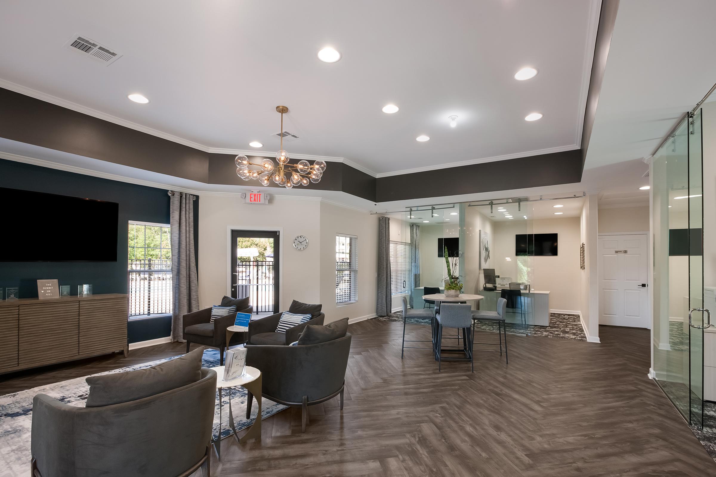 A spacious living room with modern decor, featuring a blue accent wall, two armchairs, a sofa, and a coffee table. A desk with a computer is in the foreground. Large windows provide natural light, and sliding glass doors lead to an outdoor area with greenery. The flooring is tiled.